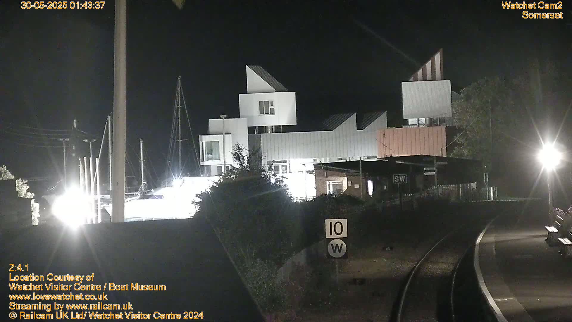 A nighttime view of a modern building complex near a marina and railway tracks, illuminated by artificial lights.
