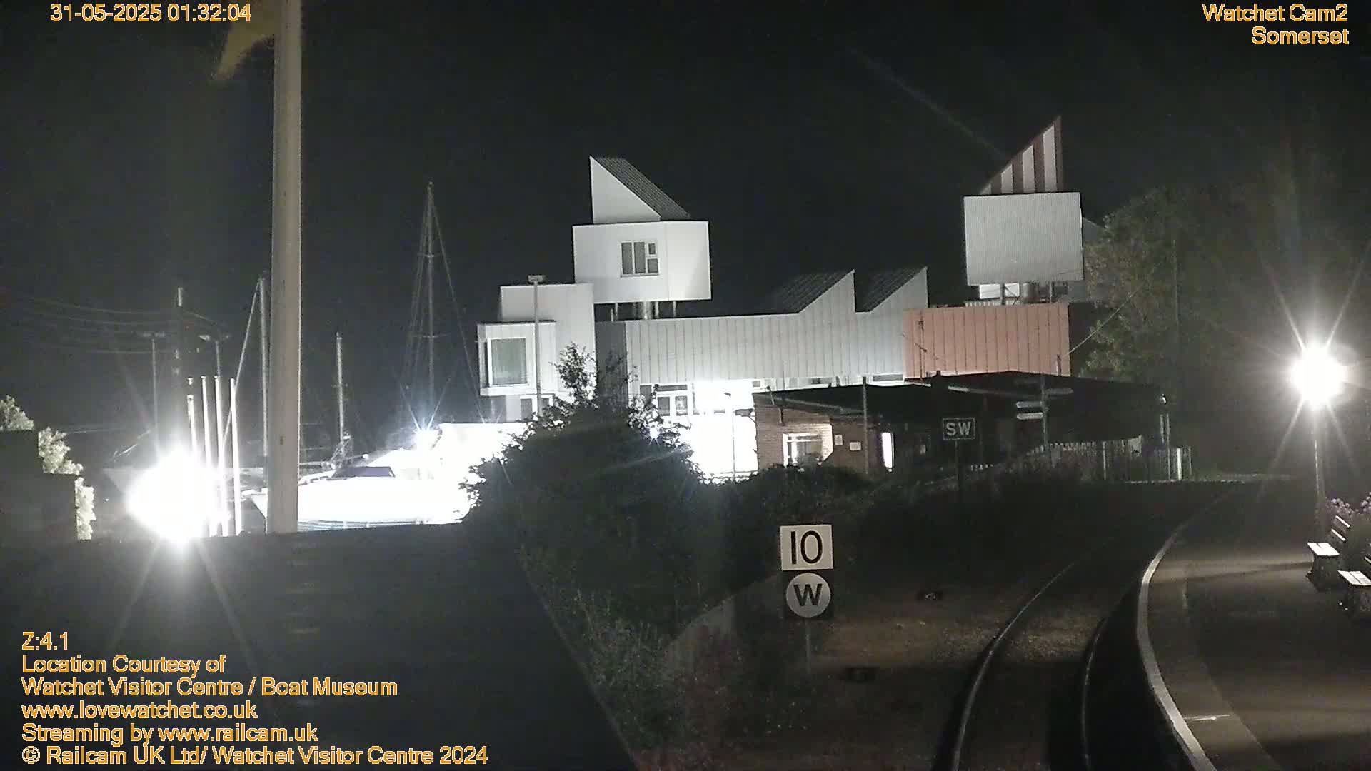 A modern, multi-level building is illuminated at night near a railway track and a boat in a marina.