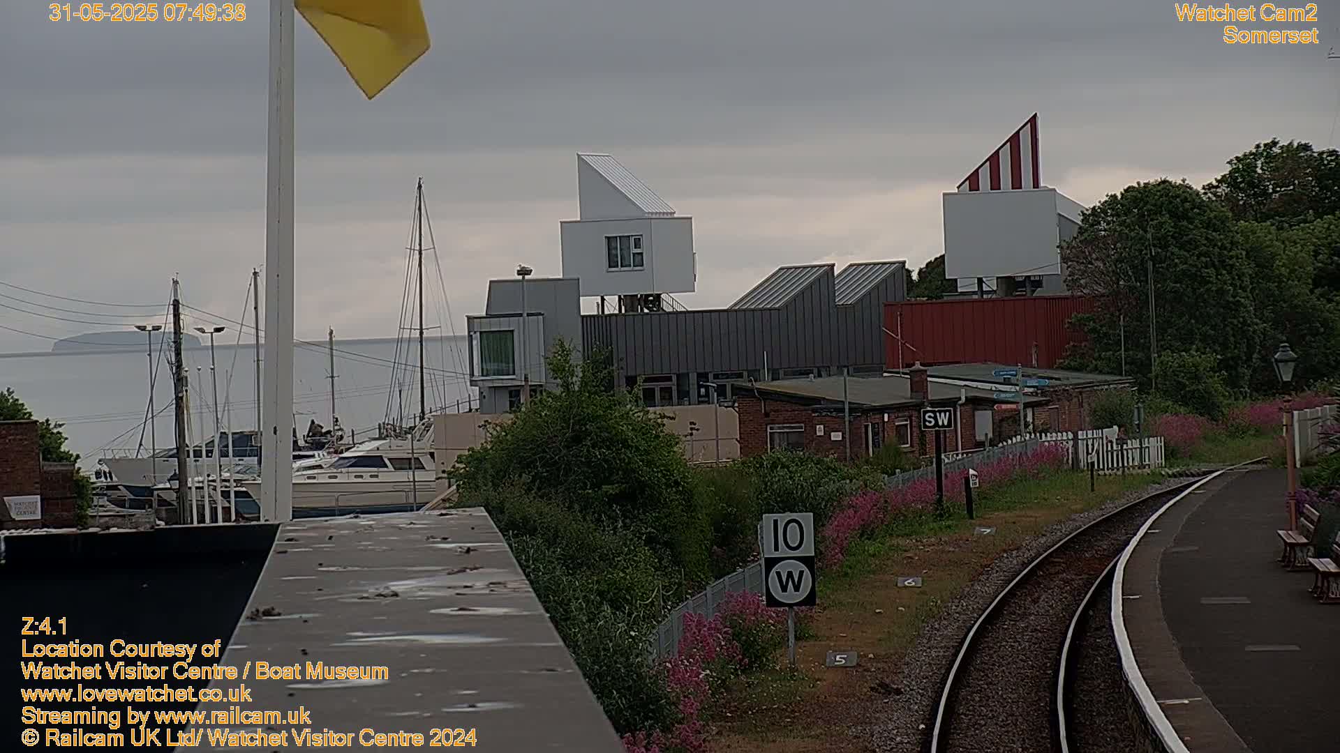A waterfront scene on an overcast day shows a marina with boats, modern buildings, and a railway line with a platform next to flowering plants.