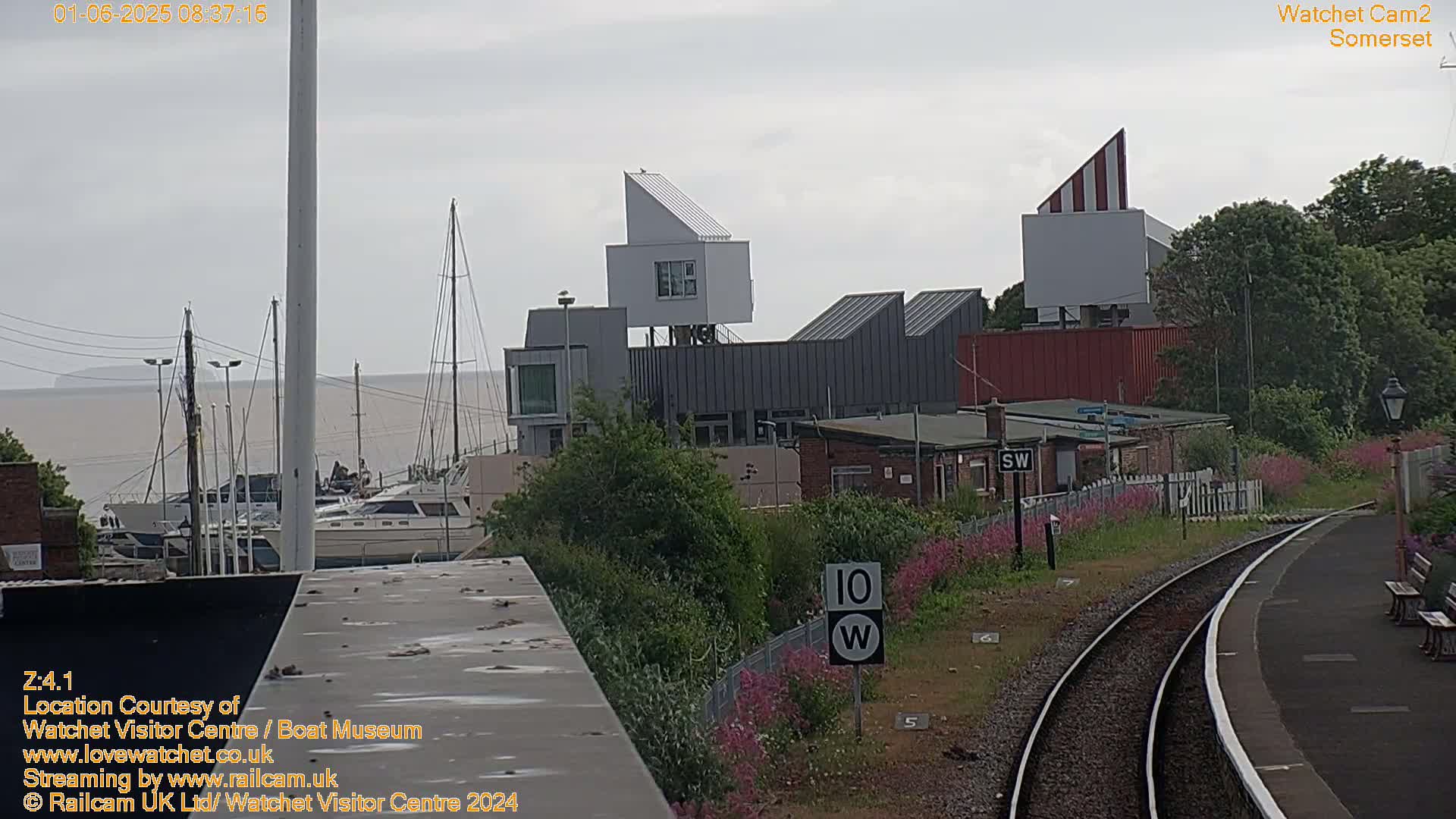 A view of a harbor with boats, modern white buildings, and a railway track under an overcast sky.