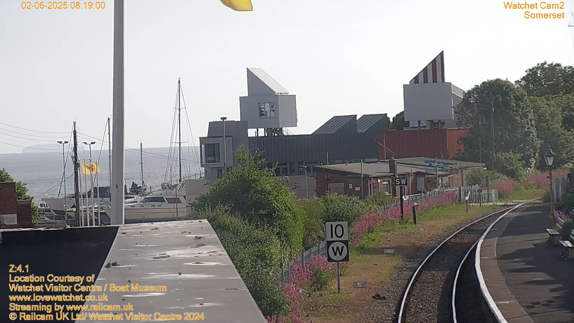 A hazy, sunny day overlooking a harbor with boats, modern buildings, and a railway track with pink flowers along the side.