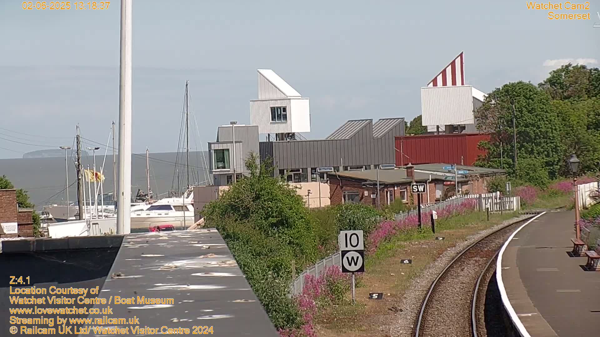 A waterfront scene on a sunny day shows a marina with boats, modern buildings, and a railway track curving toward the viewer.