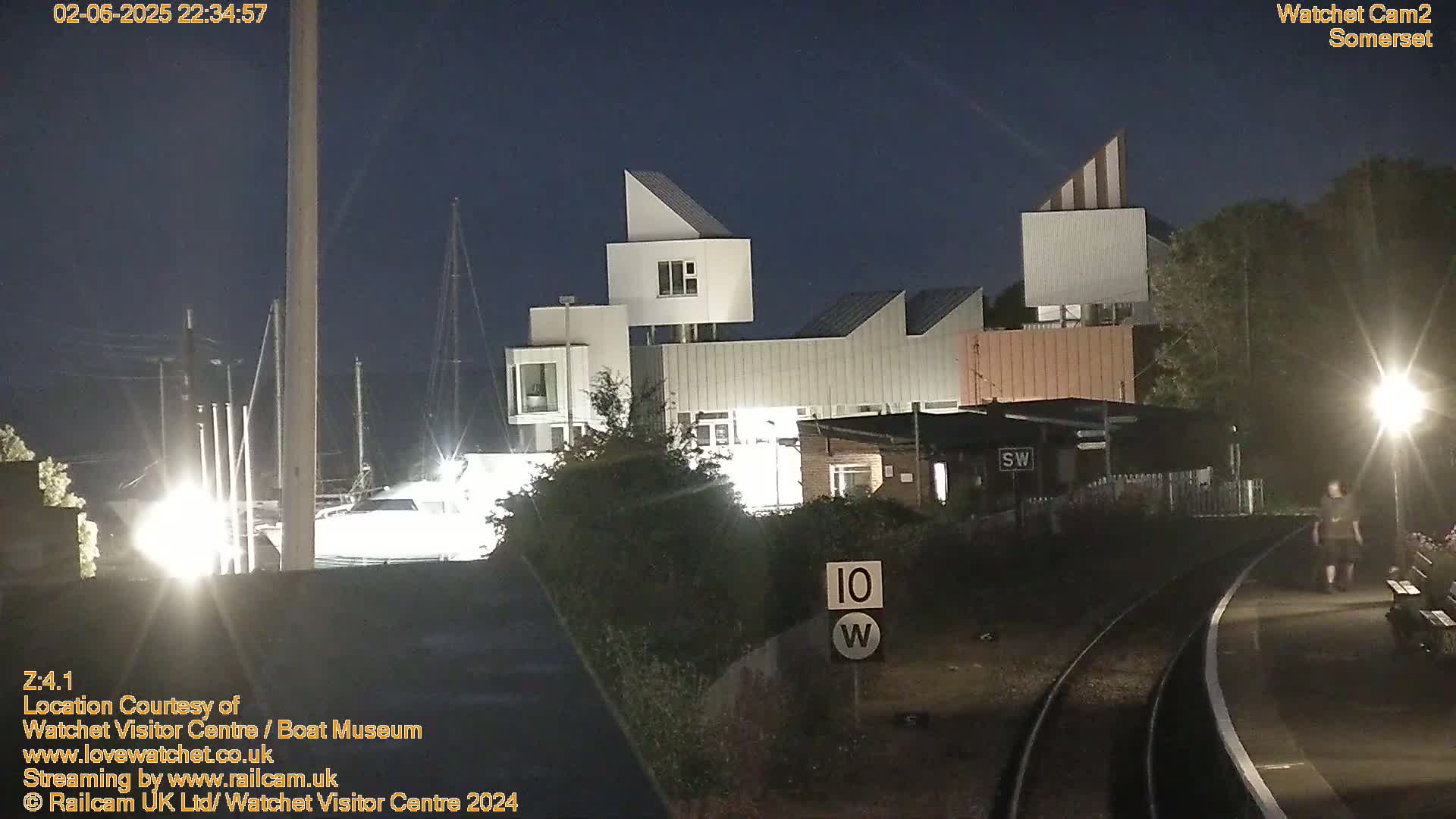 At night, a modern building complex is illuminated near a train track where a person walks, under a dark sky.