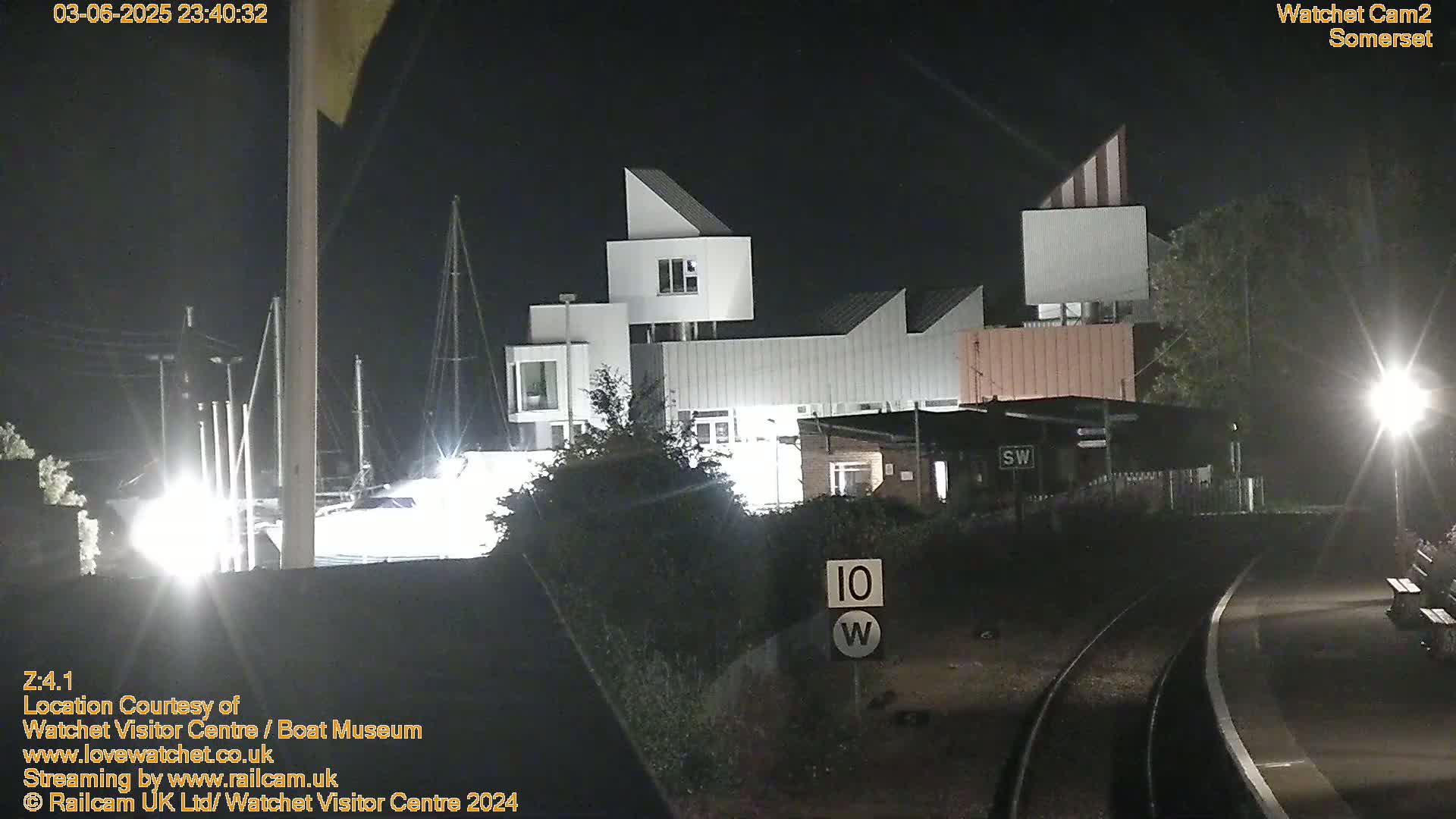 At night, a modern, white building complex is illuminated near a railway track with a speed limit sign.