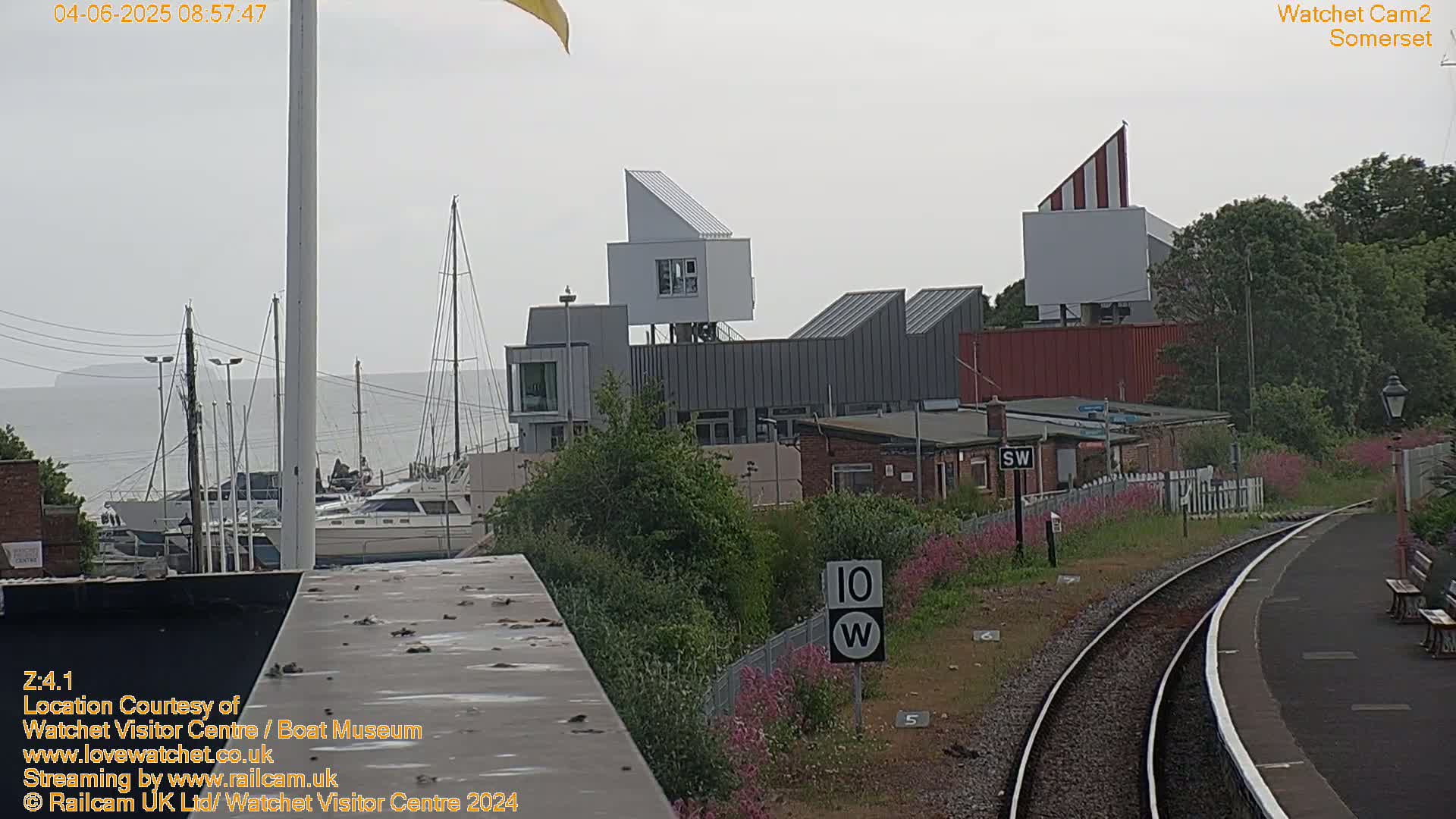 A hazy view of a harbor with boats, modern buildings, and a railway track with wildflowers alongside.