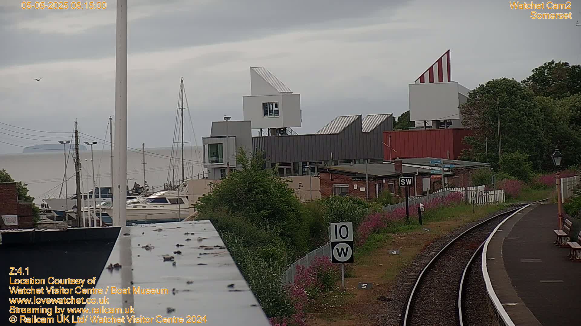 A harbor with moored boats is visible beyond a train station and modern buildings under an overcast sky.