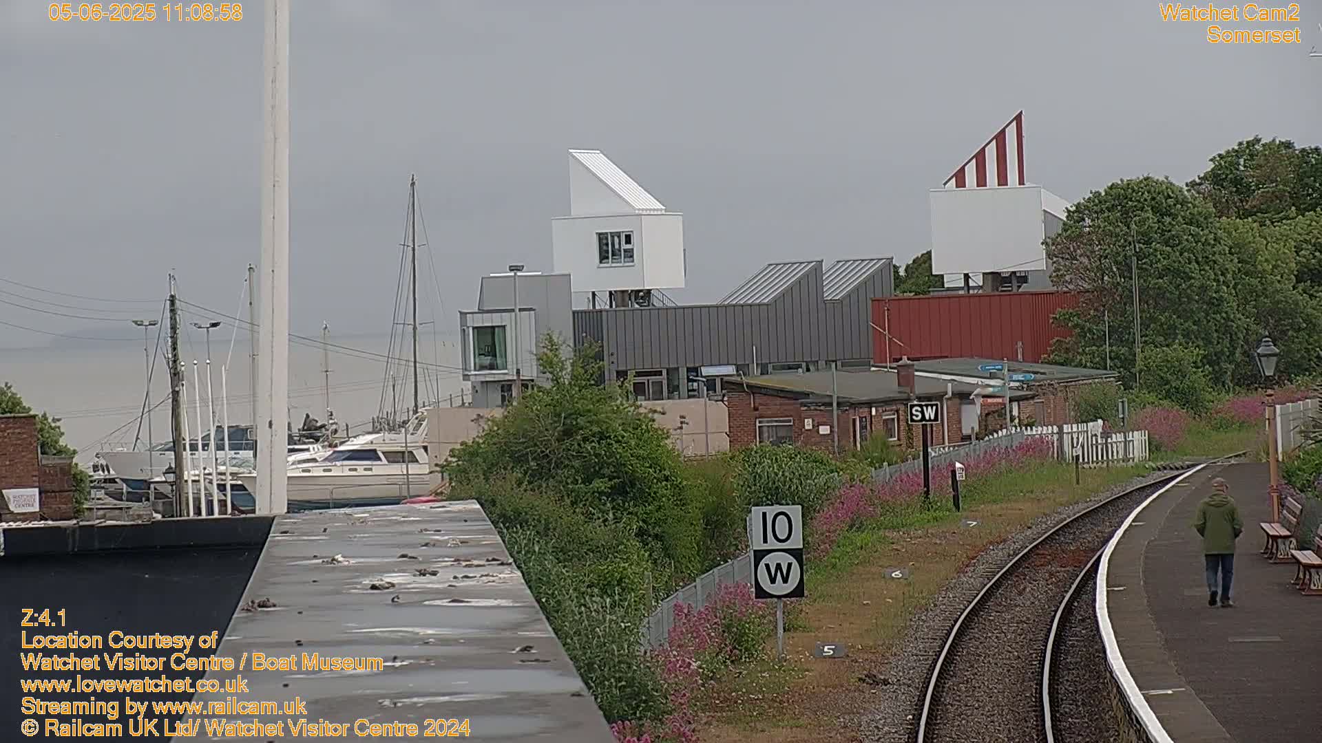 A person walks along a train platform overlooking a harbor with boats and modern buildings, under an overcast sky.