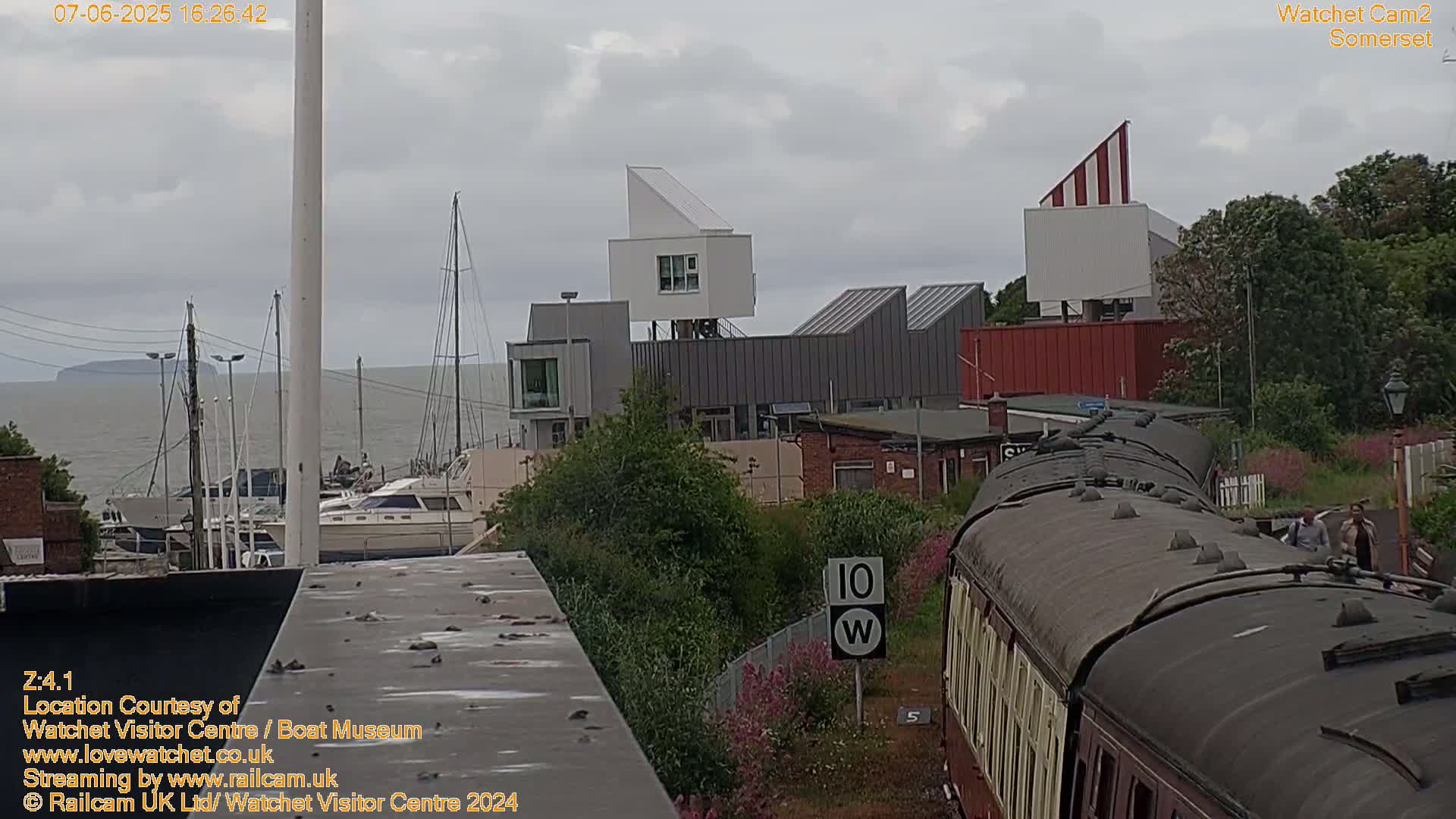 A view from a train overlooking a harbor with moored boats, modern buildings, and a cloudy sky.