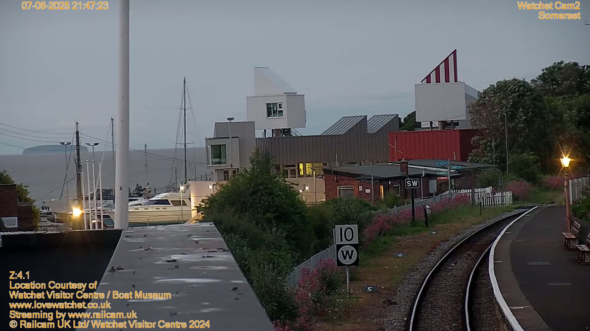 Under an overcast evening sky, a harbor scene features boats docked near modern buildings, railway tracks, and lush vegetation.