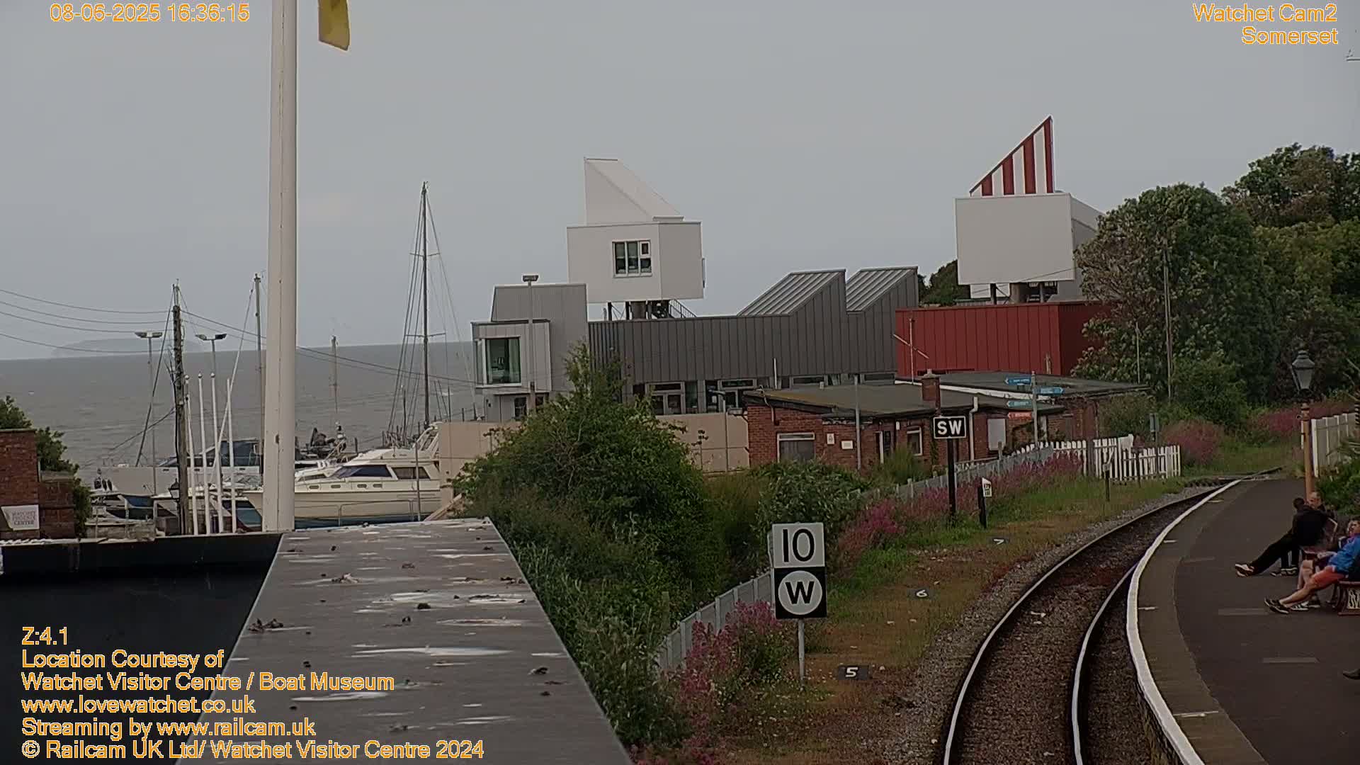 A view of a harbor with boats, modern buildings, and a train track, under an overcast sky.