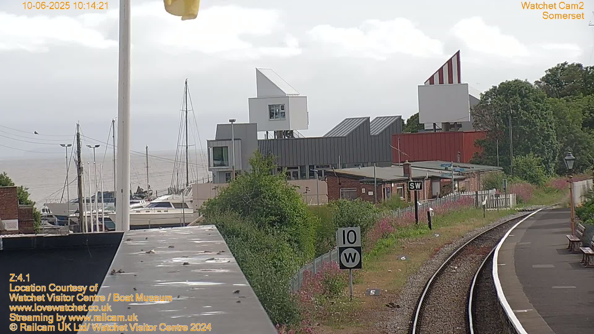 Under a cloudy sky, a harbor with moored sailboats is visible beyond a railway platform, next to modern buildings and a speed limit sign.