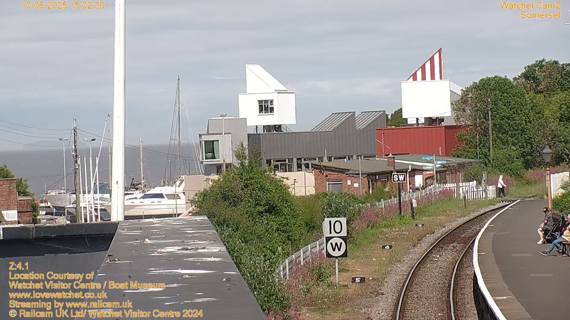 A marina with moored boats is visible beyond a modern building complex near a railway track on a partly sunny day.