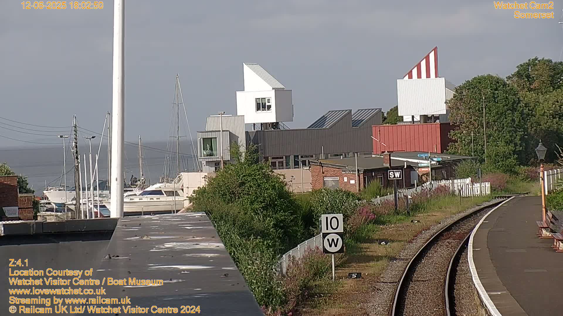 A partly cloudy day reveals a harbor with moored boats, modern buildings, and a railway track with a speed limit sign.