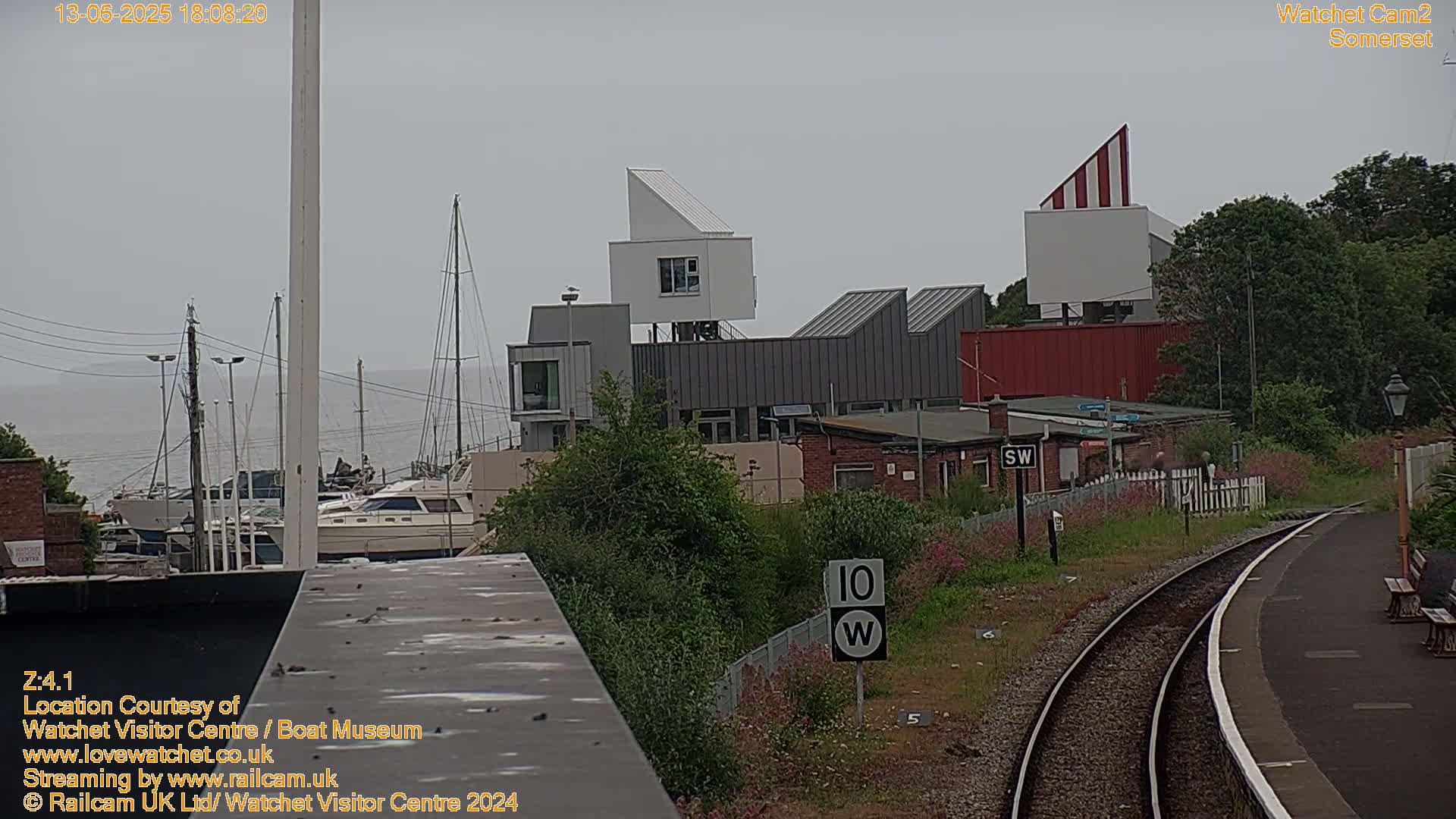 A marina with boats, modern white buildings, and a railway line is visible under an overcast sky.