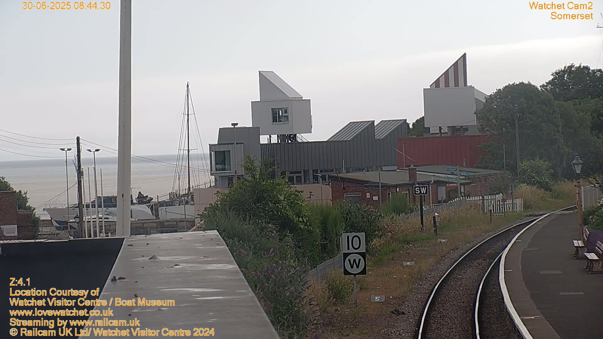A view of a harbor with boats, a modern building complex, and a railway track under an overcast sky.