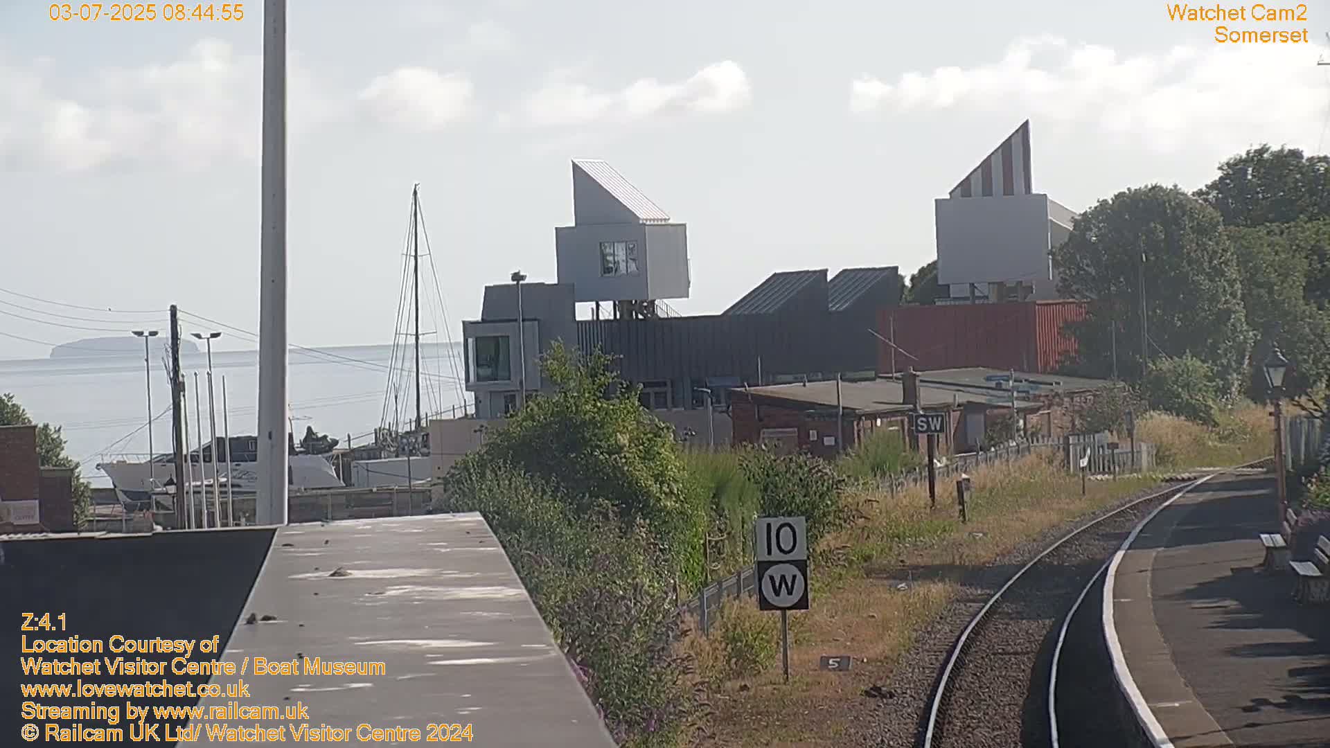 A waterfront scene on a sunny day shows modern buildings near a train station and harbor with boats, viewed from a slightly elevated vantage point.