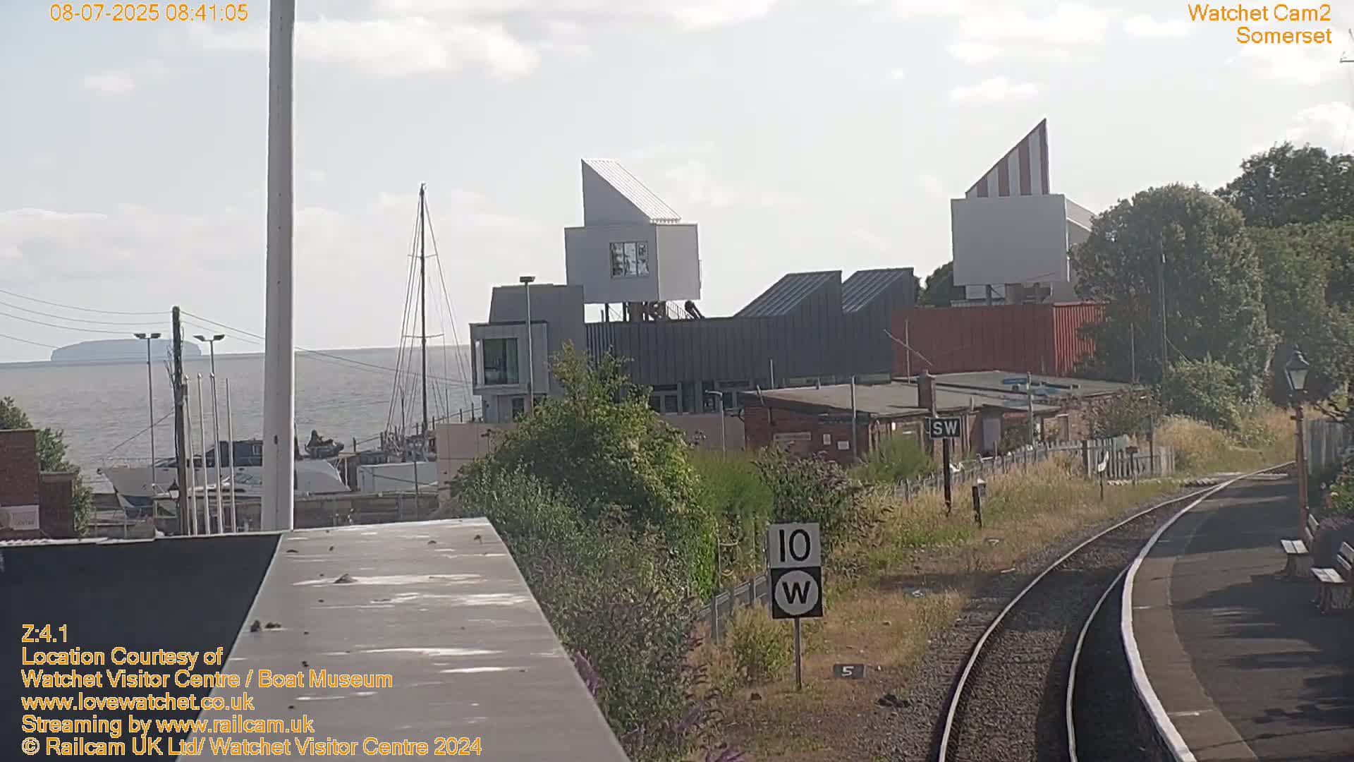 A view from a train shows a modern, white-and-gray building complex near a body of water under a partly cloudy sky.