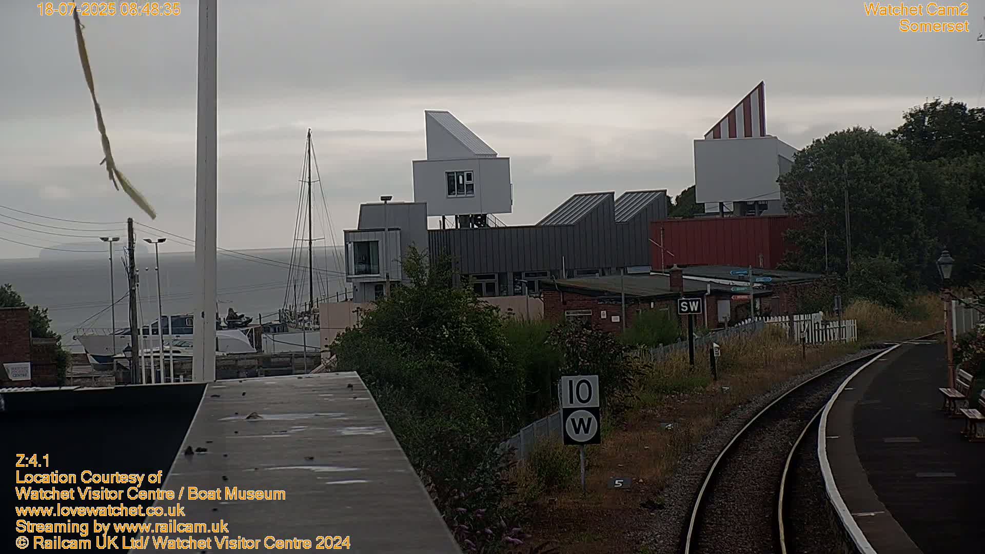 A modern, angular building complex sits near a body of water under an overcast sky, with railway tracks visible in the foreground.