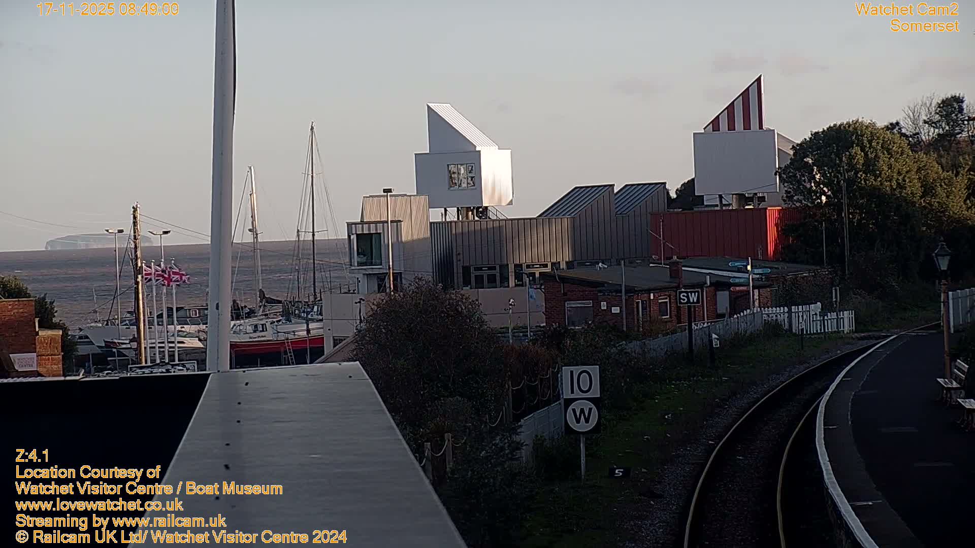 A clear sky hangs over a bustling coastal scene featuring a harbor with numerous sailboats, several modern buildings with angular designs, and a railway line with a platform curving into the foreground.