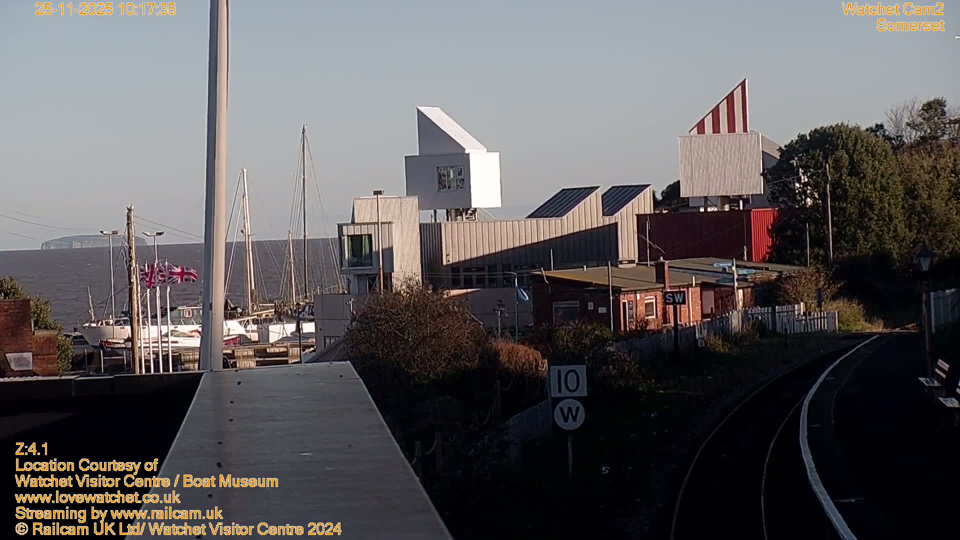 The image captures a sunny, clear day at a coastal location, featuring a harbor with moored boats and a distant island beyond modern and traditional buildings, with train tracks and a platform curving into the foreground.