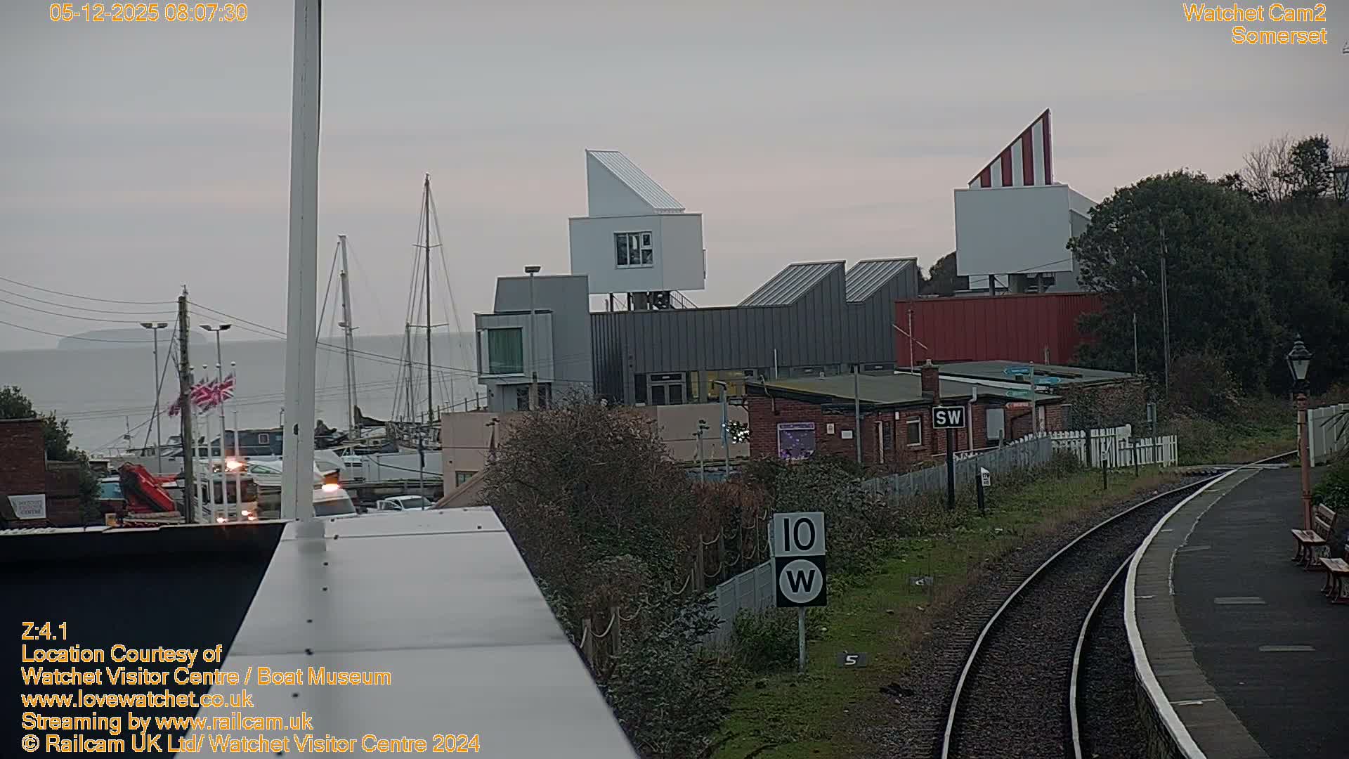 On an overcast and grey day, a curving train track and platform occupy the right side of the image, while on the left, a harbor filled with boats and several modern and industrial buildings sits against a backdrop of distant hills.
