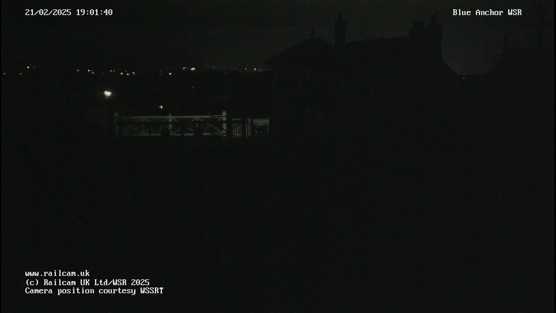 A dark nighttime image shows a dimly lit building and fence in the distance under a clear night sky.