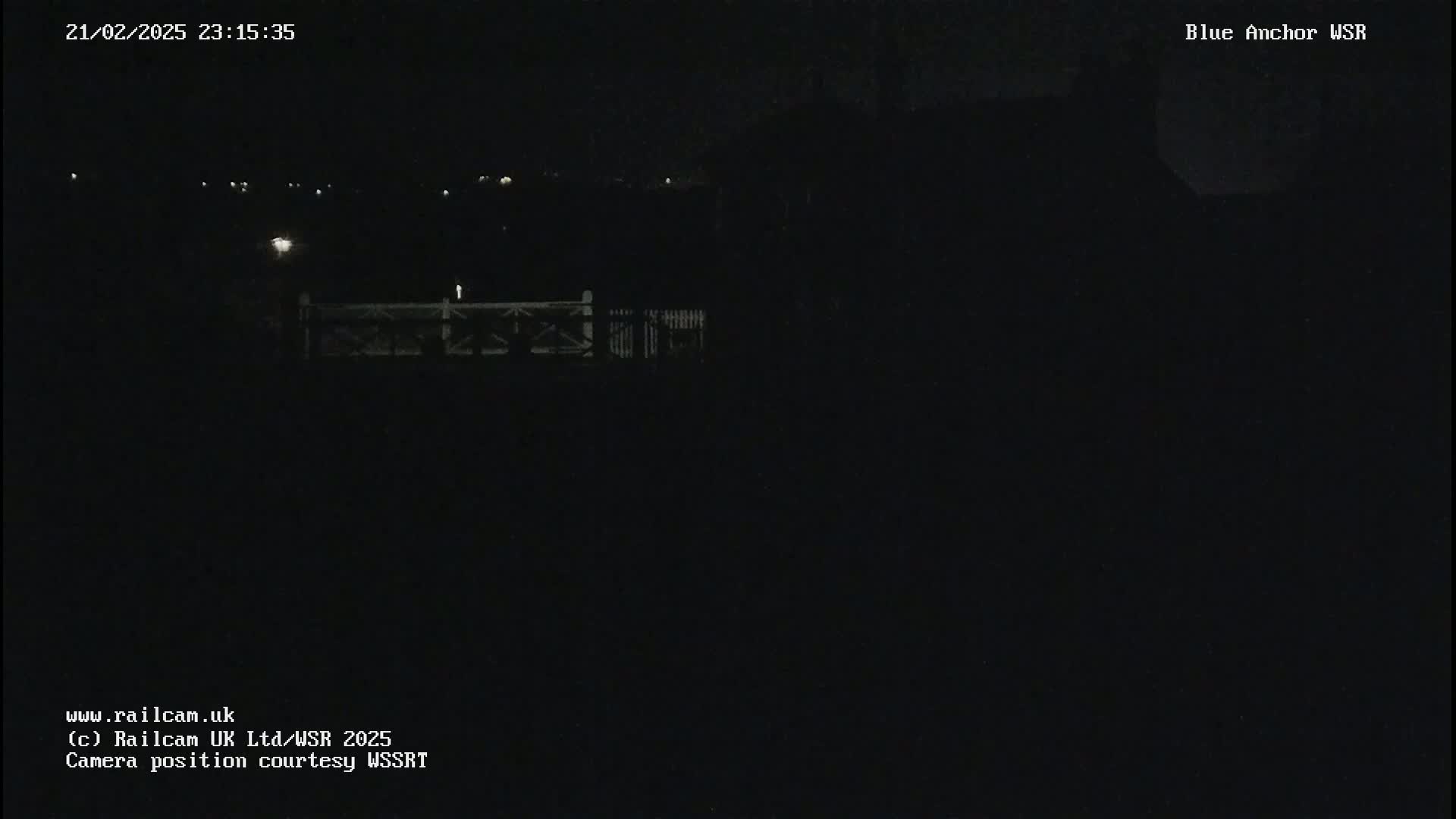 A nighttime, low-light view of a dark landscape featuring a fence and distant lights.