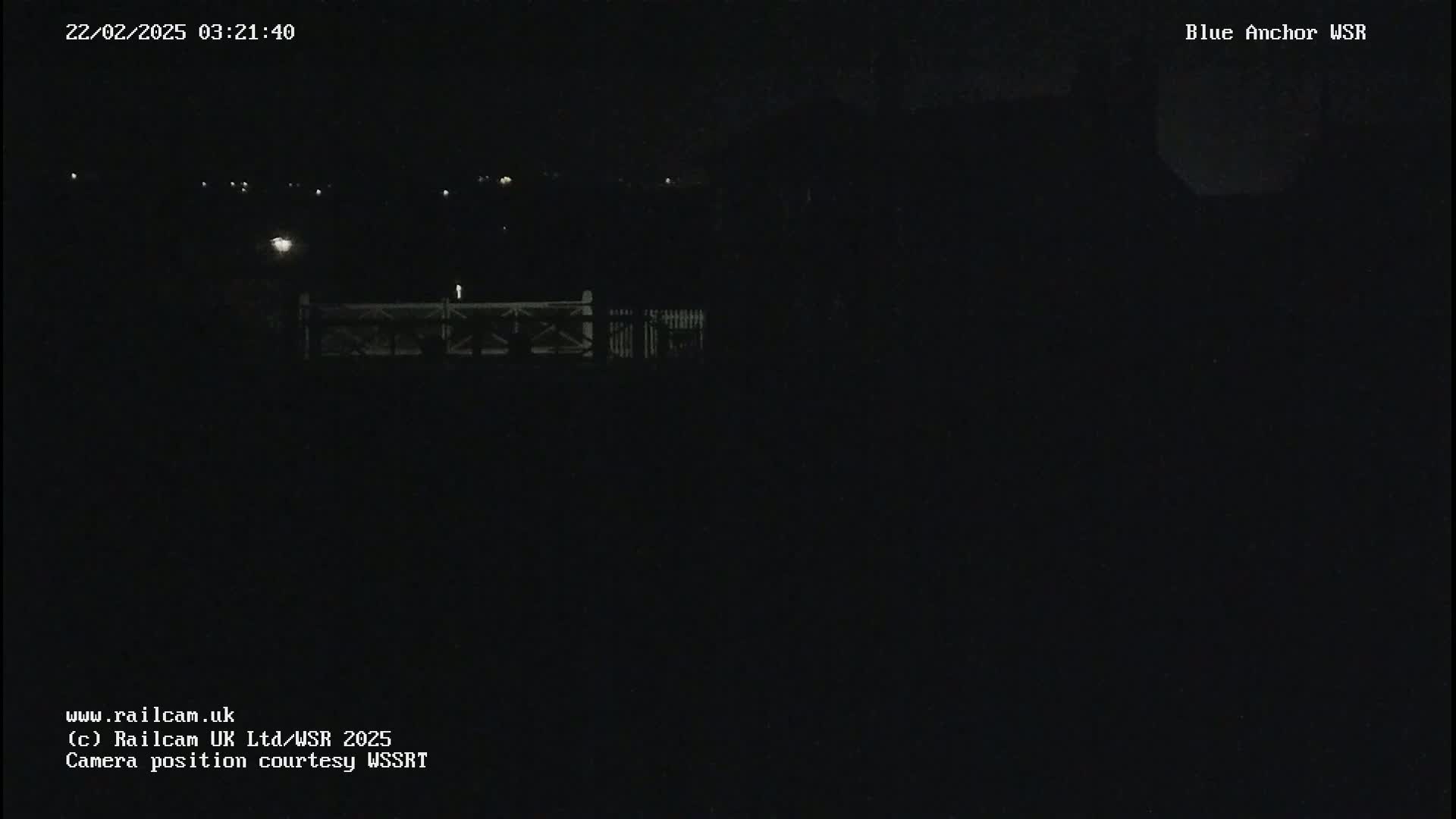 A nighttime, low-light view of a dark landscape featuring a fence and distant, faint lights.