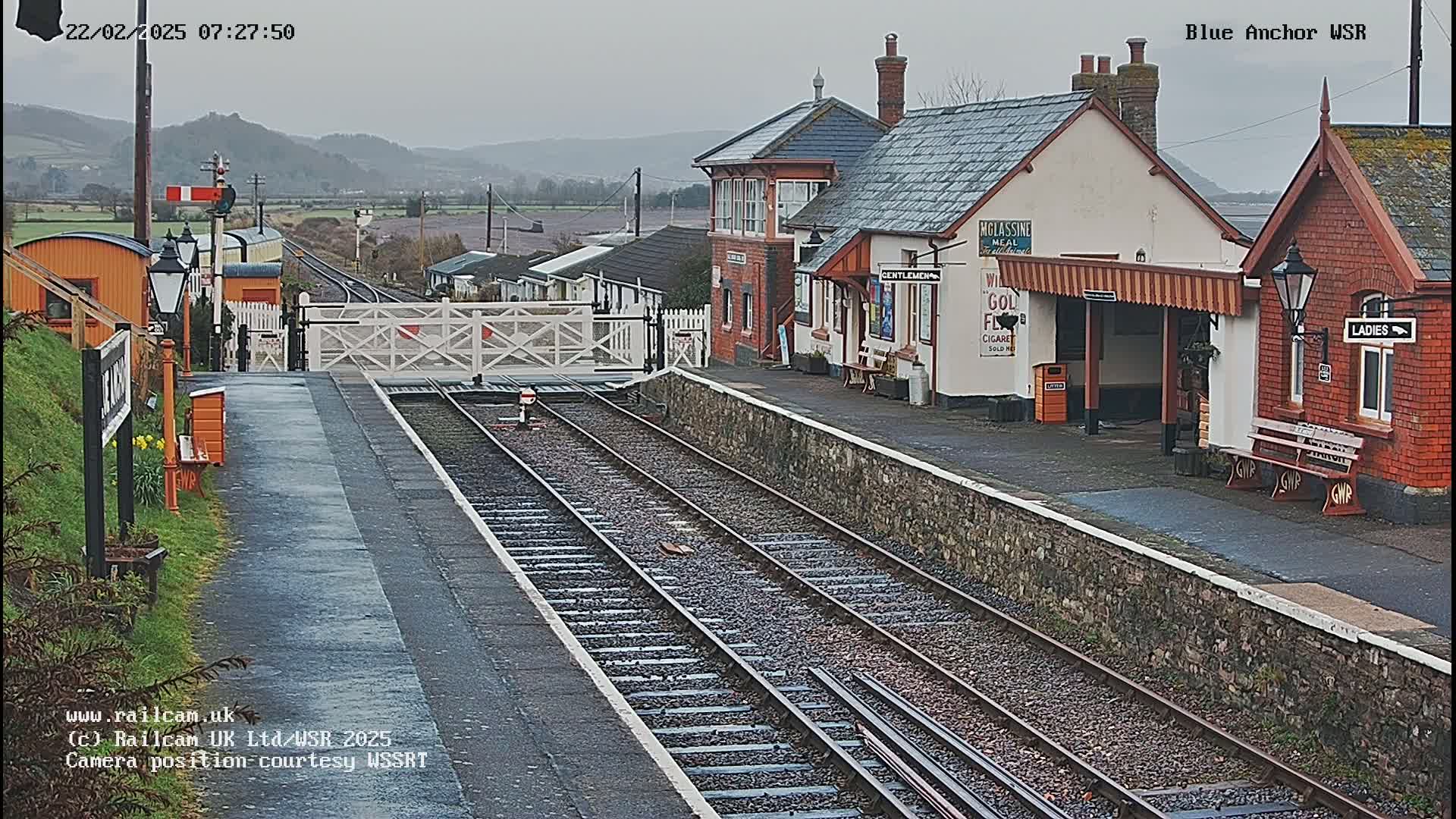 A railway station with a signal gate, train tracks, and several buildings under an overcast sky.