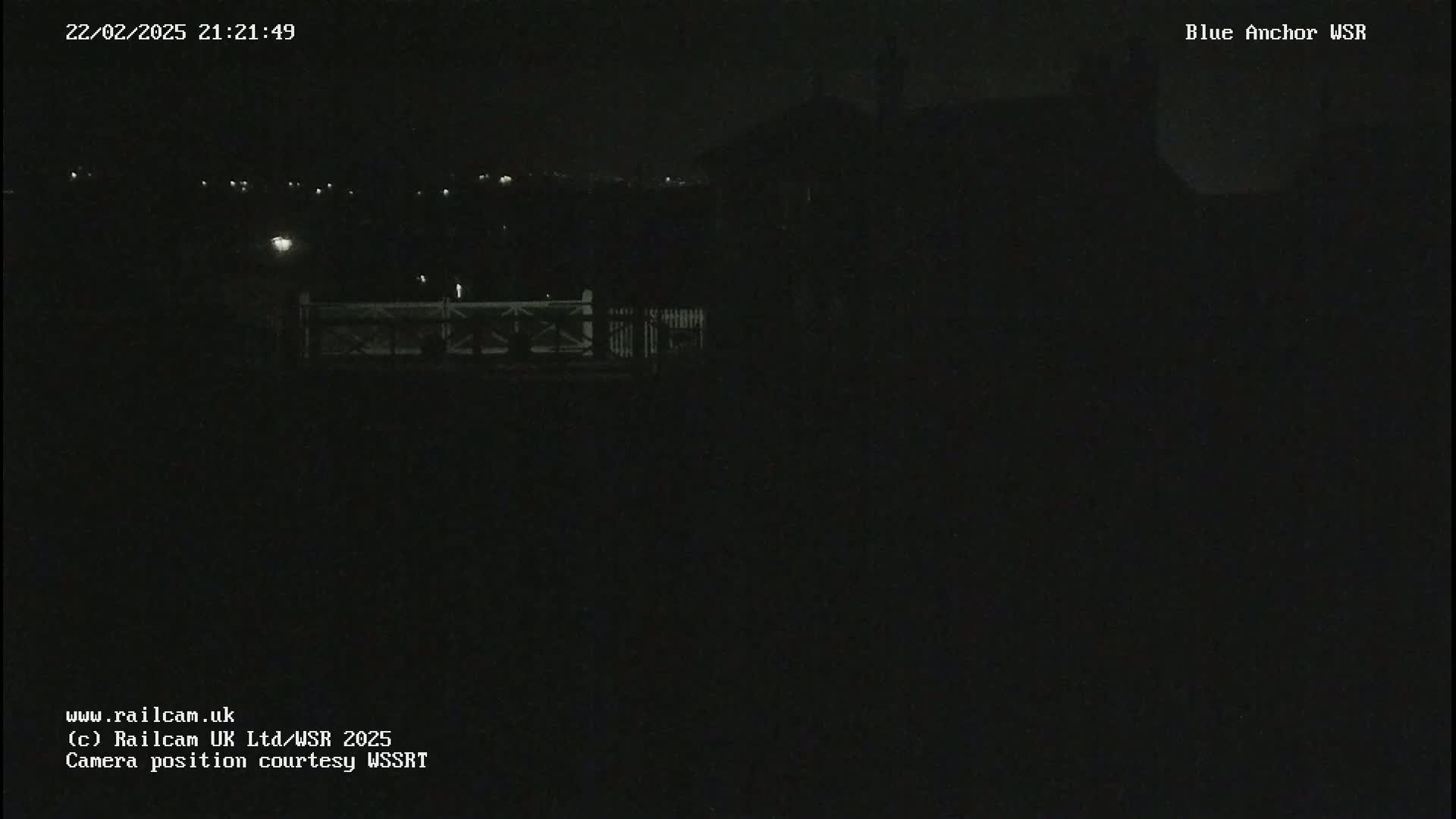A dark, nighttime scene shows a dimly lit fence and building in the distance under a clear sky.
