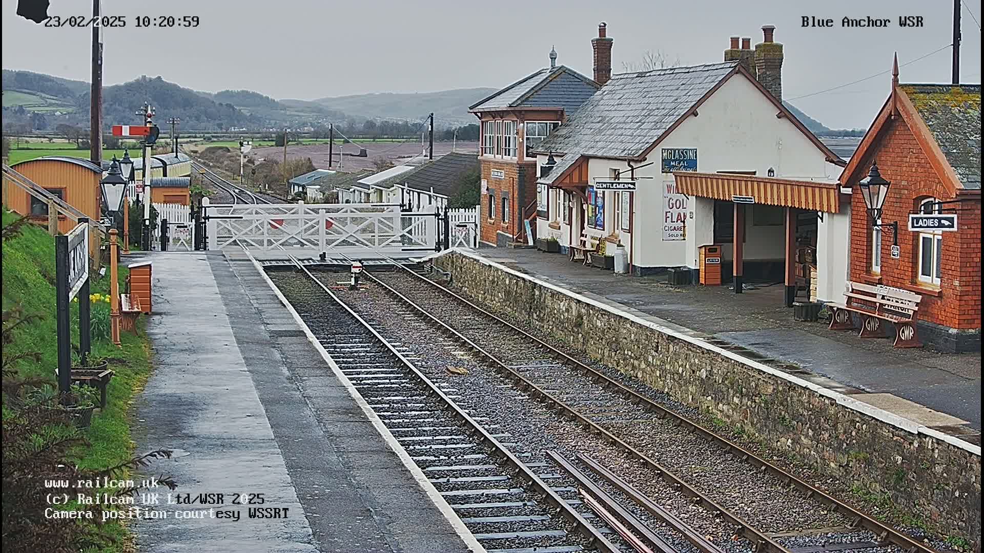 A train station with two platform buildings, a closed level crossing, and multiple train tracks under an overcast sky.