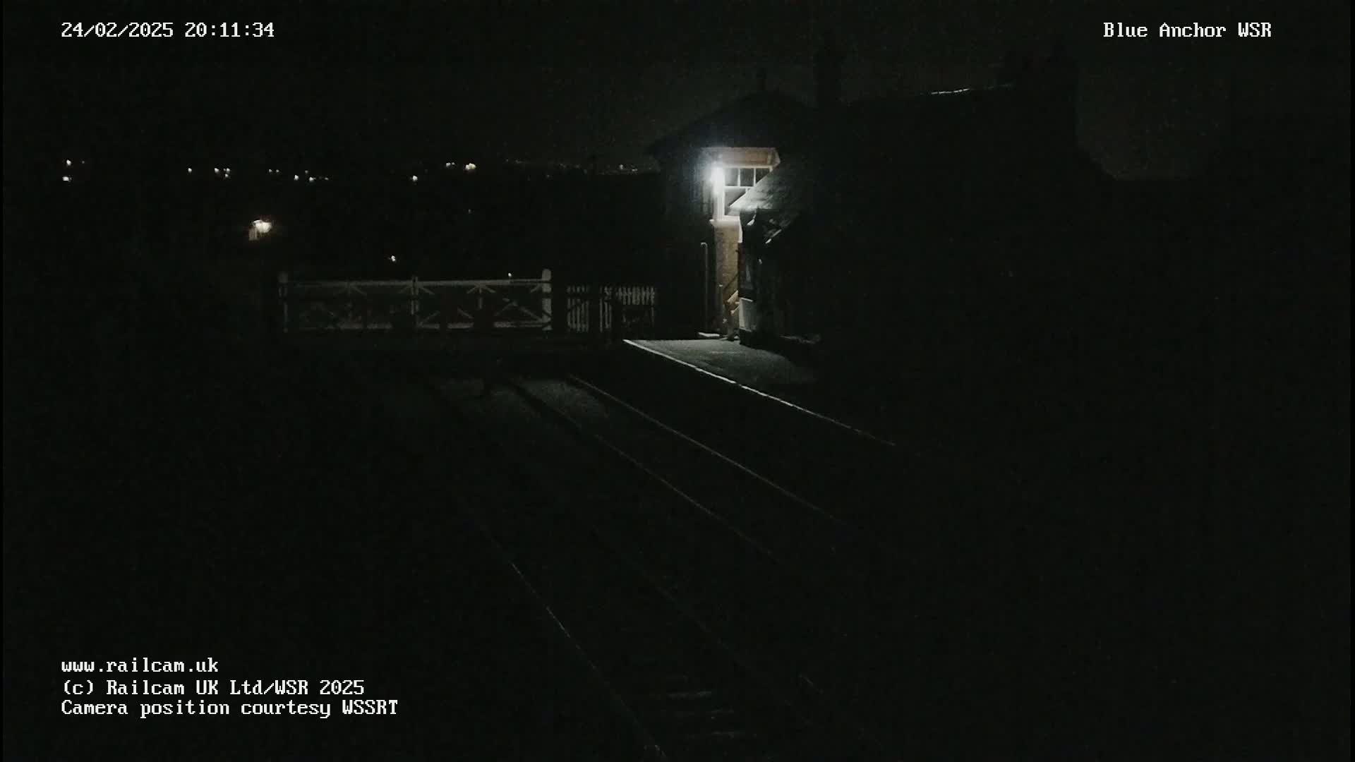 A dimly lit nighttime train station platform is shown with a building illuminated by a single light.