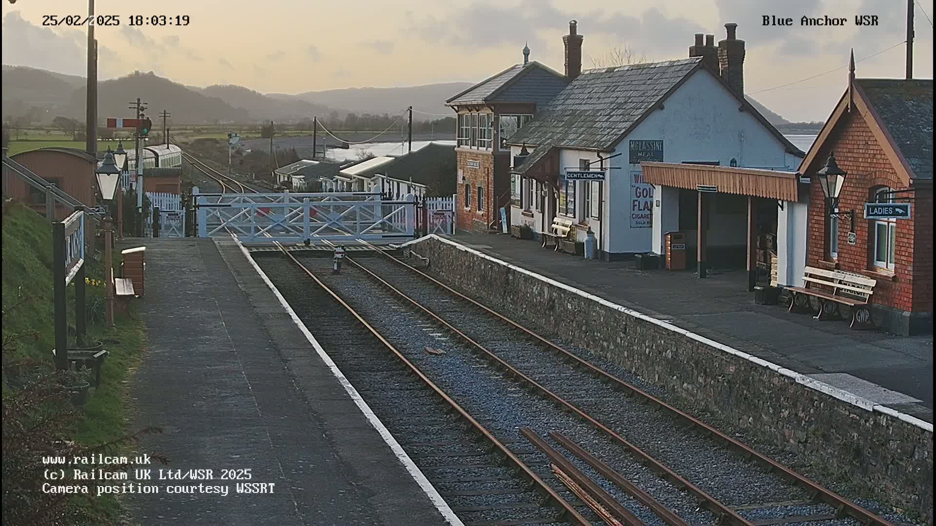 A train station with two platform buildings and a level crossing, under a partly cloudy twilight sky, with hills visible in the distance.