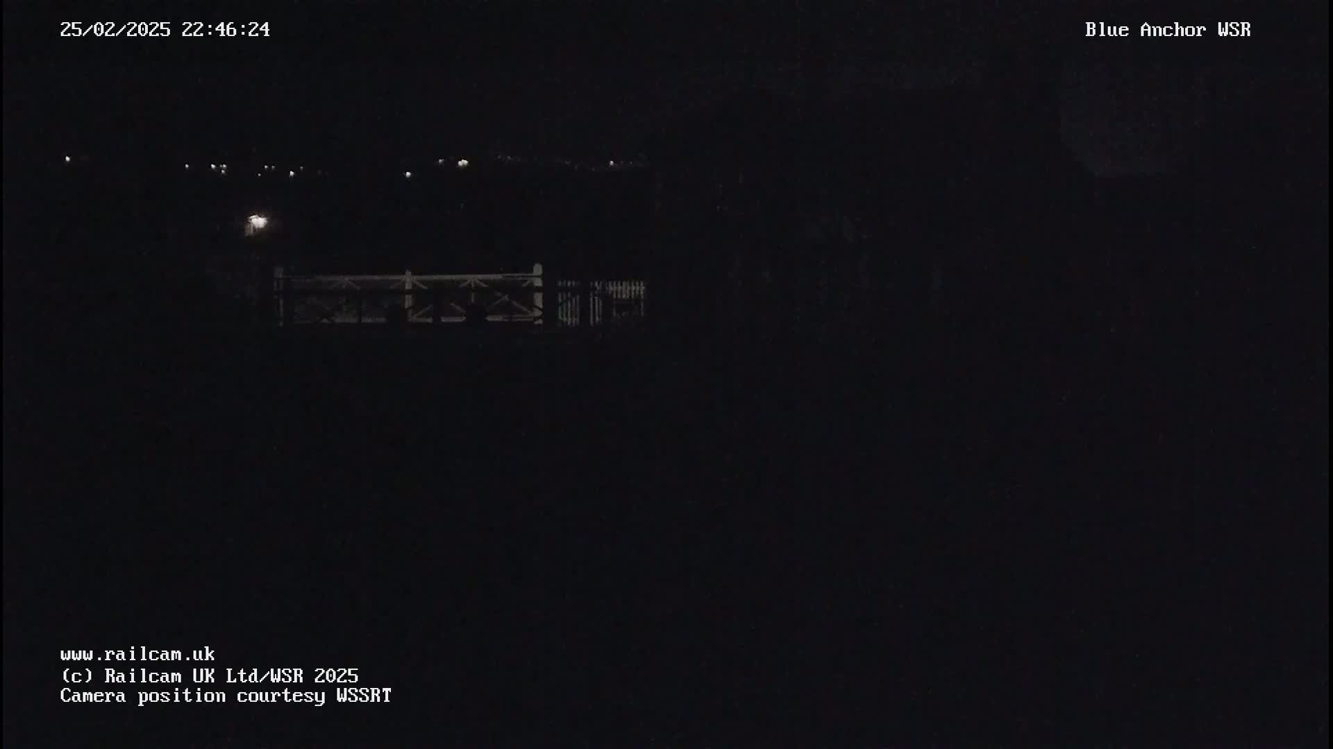 A nighttime, dark image shows a dimly lit fence and distant lights under a dark sky.