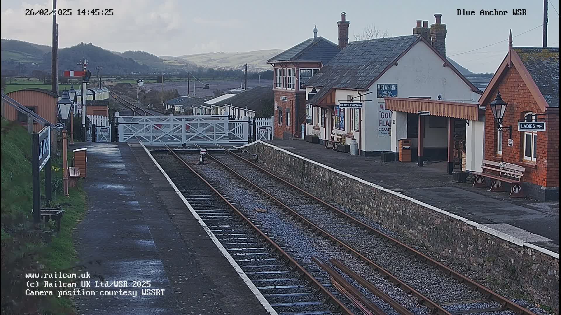 A train station with a railway crossing gate and tracks, under a partly cloudy sky, is situated in a rural area with distant hills.