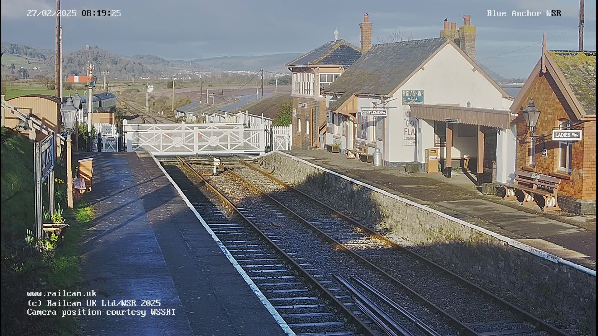 A sunny day at a train station with two parallel sets of tracks, a station building, and a gate.