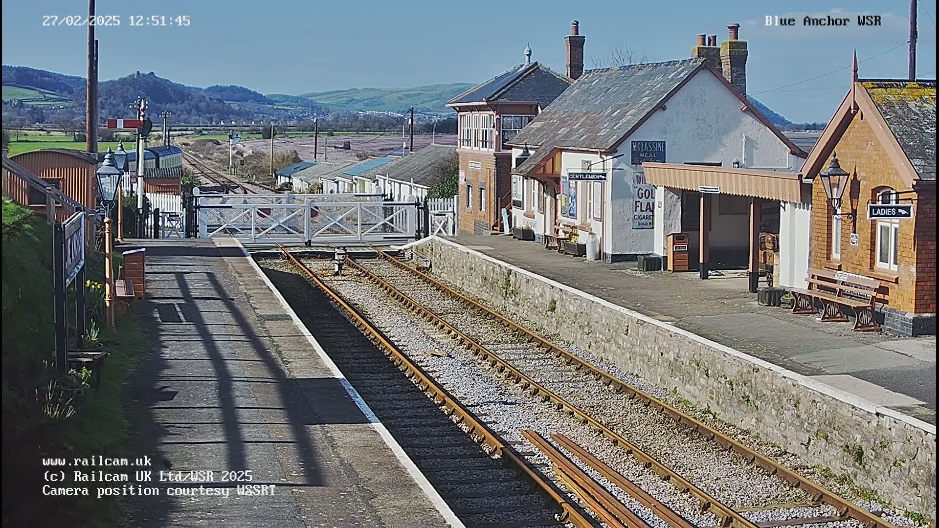 A sunny day at a train station with multiple train tracks, a signal box, and several small buildings.