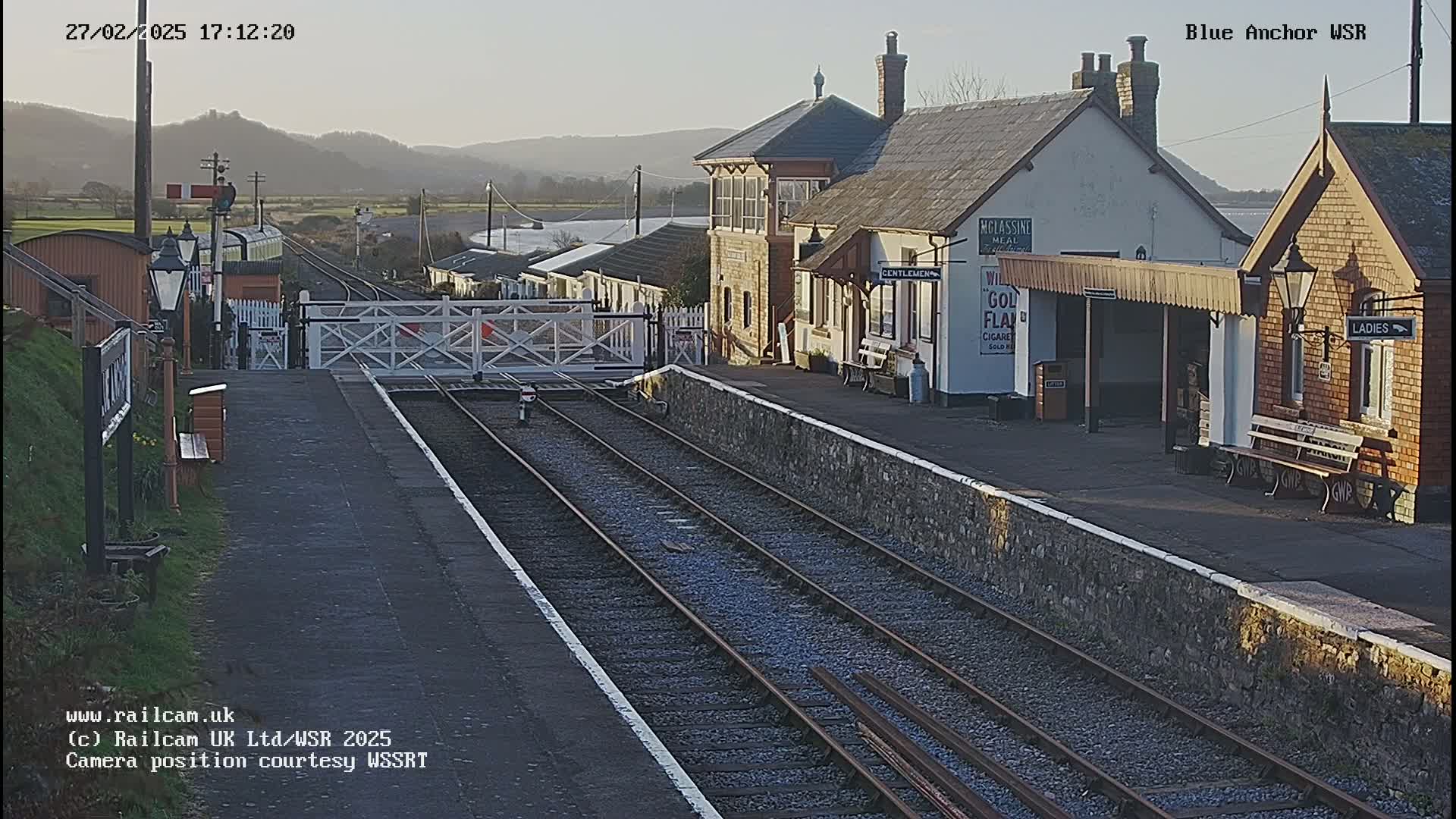 A train station with a railway track, signal crossing gates, and several buildings is visible under a clear, sunny sky.