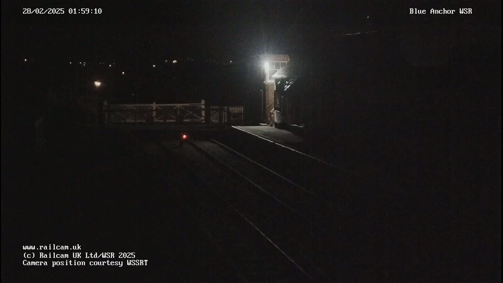 A dark, nighttime view of a train station platform with train tracks and a single light illuminating a portion of the scene.