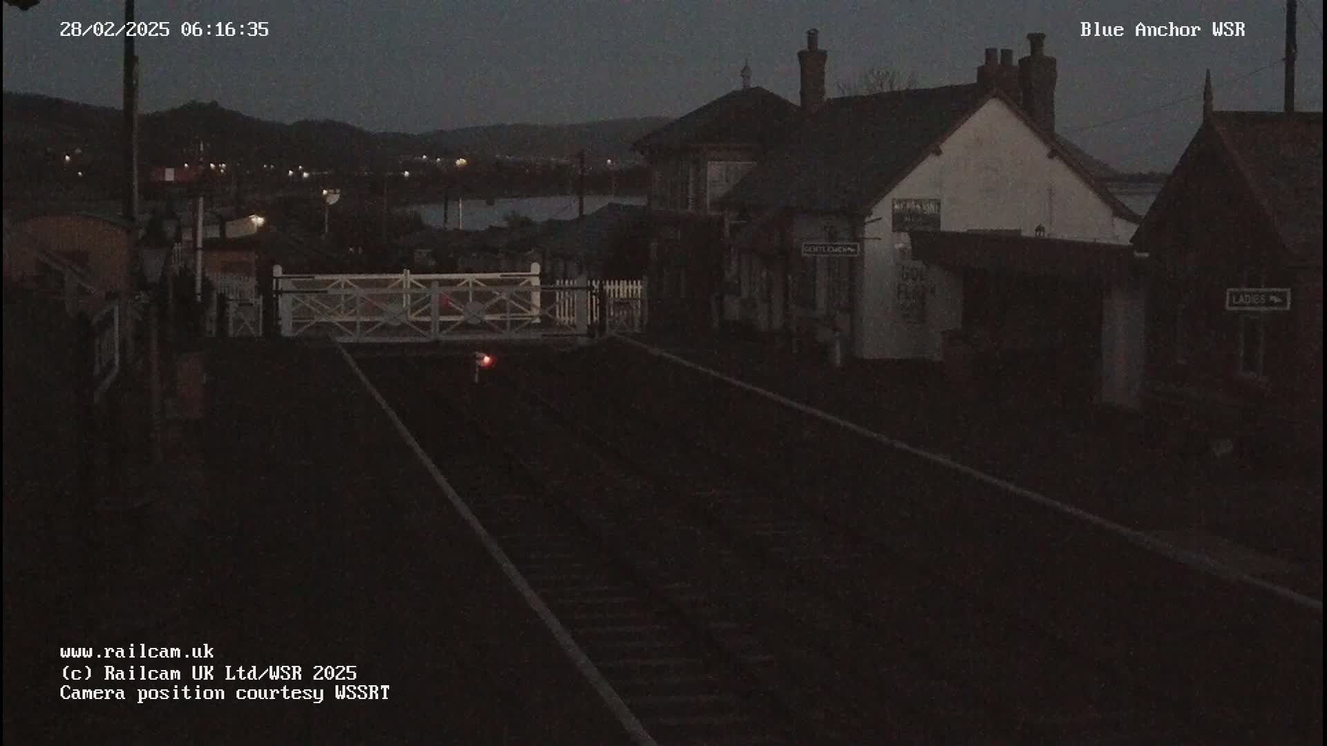 A dark, nighttime view of a railway station with a closed level crossing and train tracks extending into the distance.