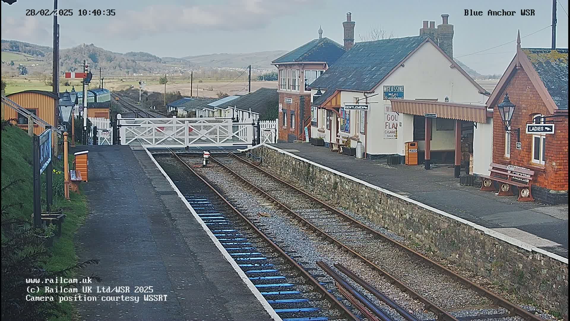 A train station with several buildings, a waiting area, and train tracks, on a partly sunny day with distant hills visible in the background.