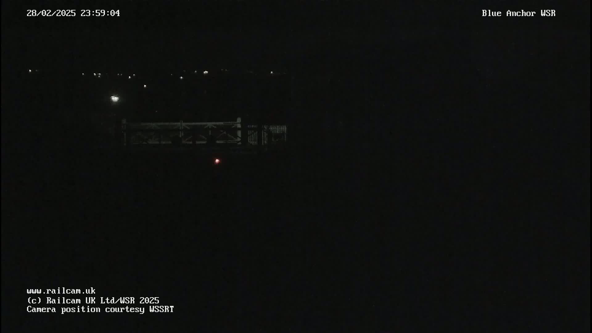 A nighttime, dark scene shows a dimly lit bridge with faint lights in the distance under a clear, dark sky.