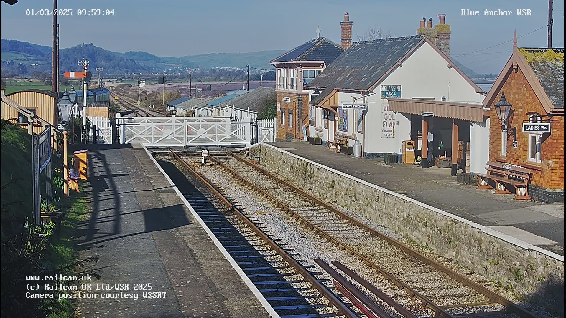 A sunny view of a train station with two sets of train tracks, a low stone wall, and station buildings under a clear blue sky.