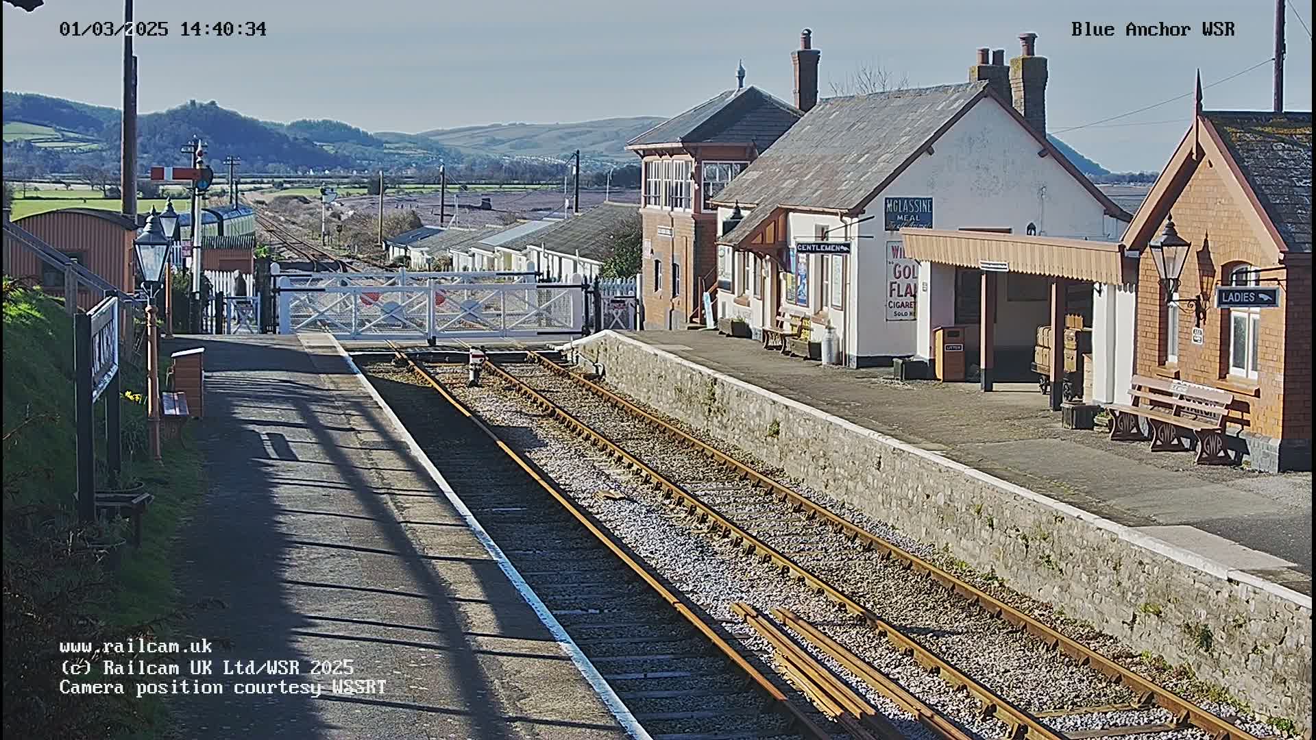 A sunny day at a train station with multiple railway tracks, a signal box, and several buildings, including a waiting area with benches.