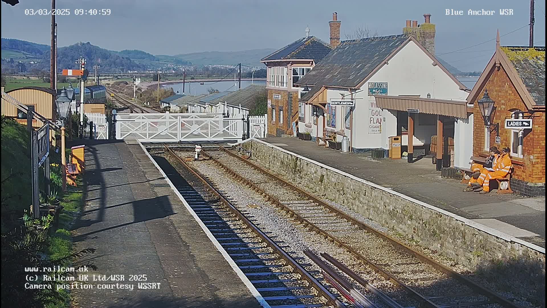 A sunny day at a train station with two people sitting on a bench near a brick building and train tracks extending into the distance.