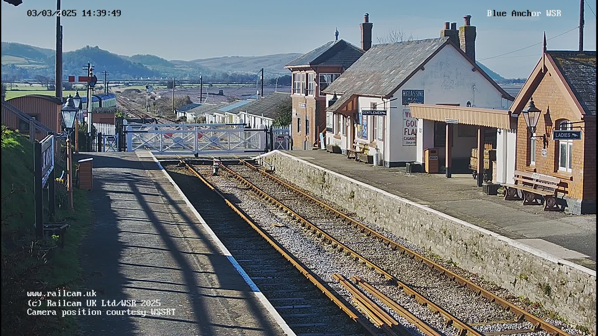 A sunny day at a train station with a signal gate down, multiple train tracks extending into the distance, and several low brick buildings.