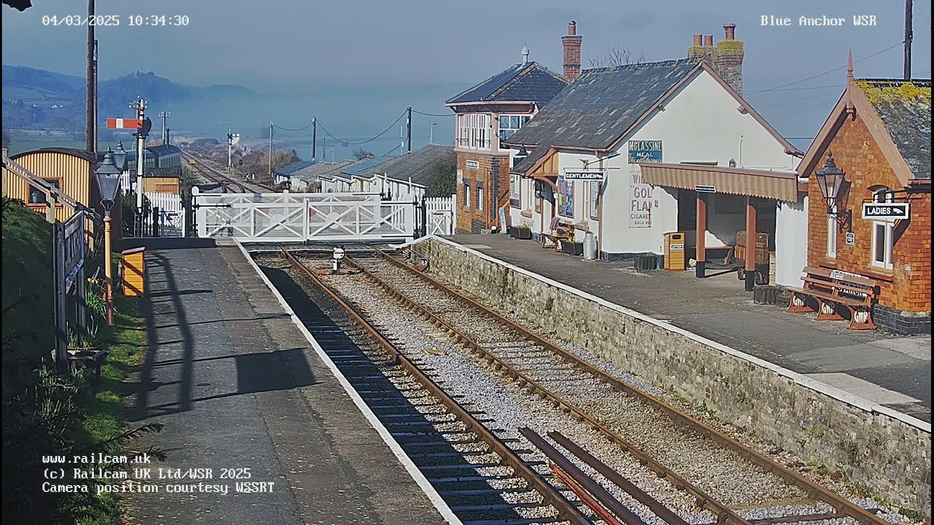 A sunny view of a train station with two sets of tracks, a signal, and several small buildings.