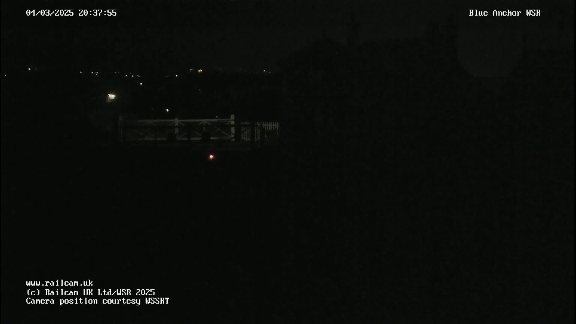 A nighttime, low-light view shows a dark landscape with a faintly visible bridge and a few distant lights.