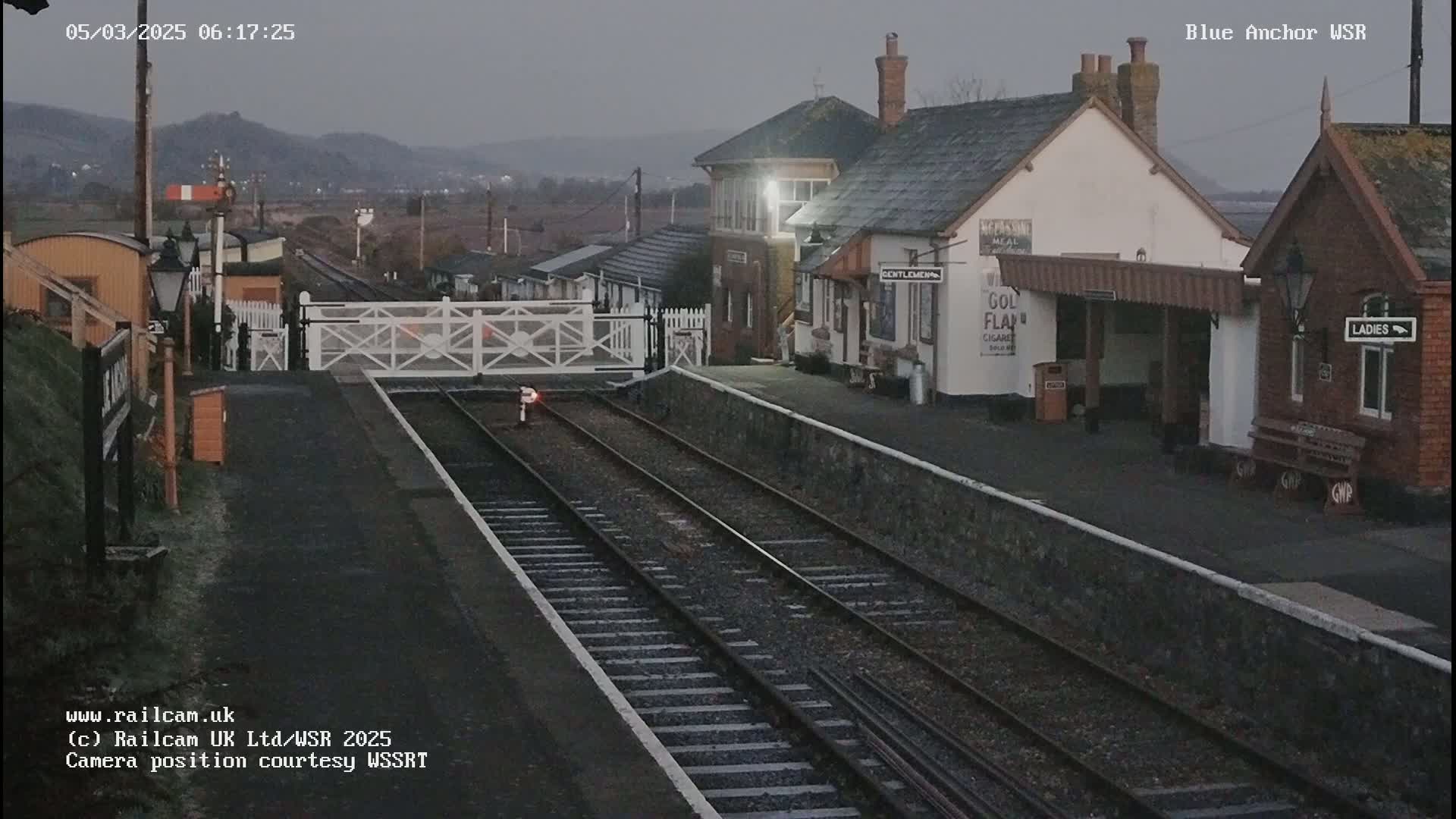 A train station with two parallel train tracks, a signal gate, and station buildings under an overcast sky.