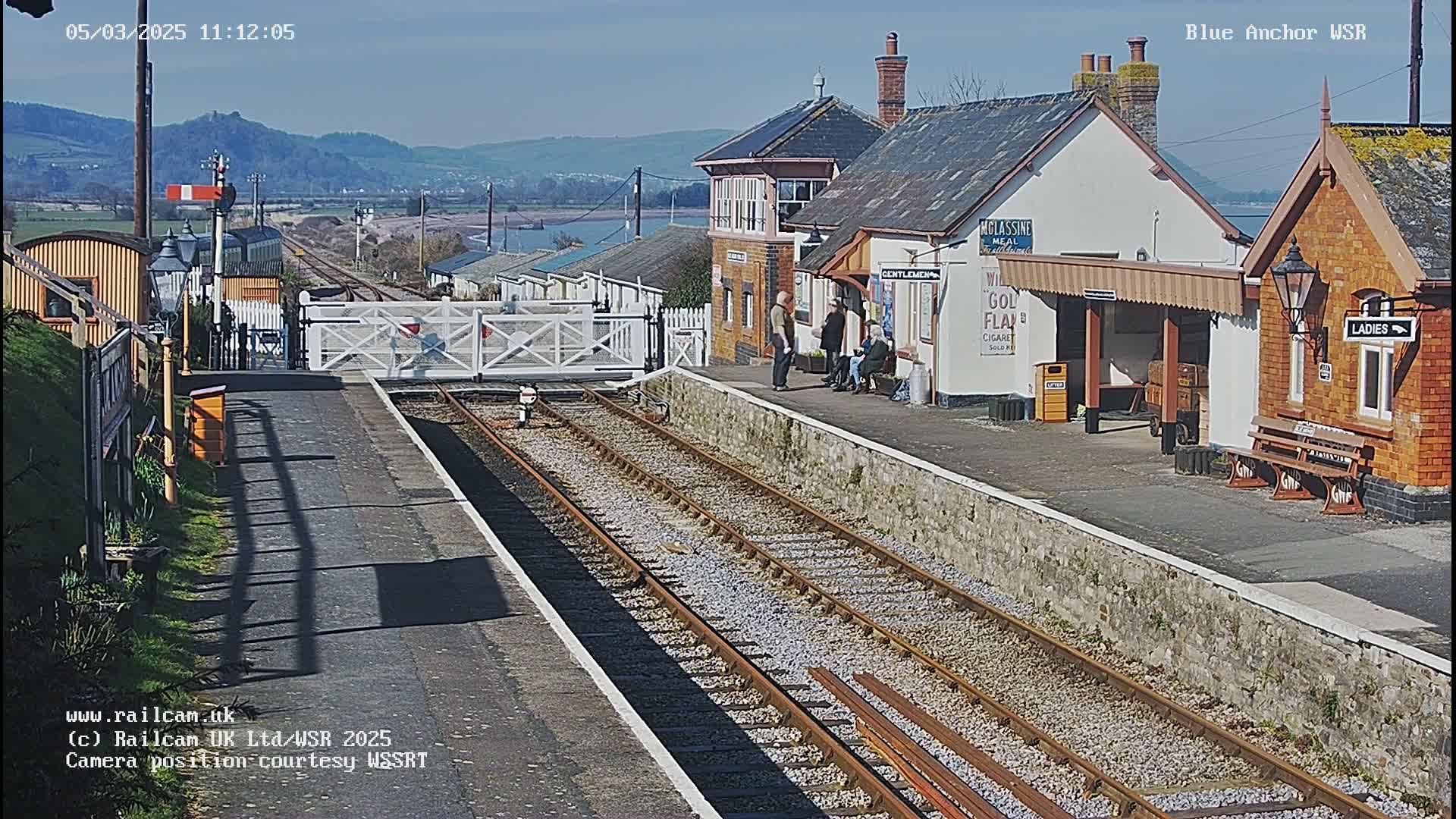 A sunny day at a train station with a few people standing near brick buildings and a train track extending into the distance, with mountains visible in the background.
